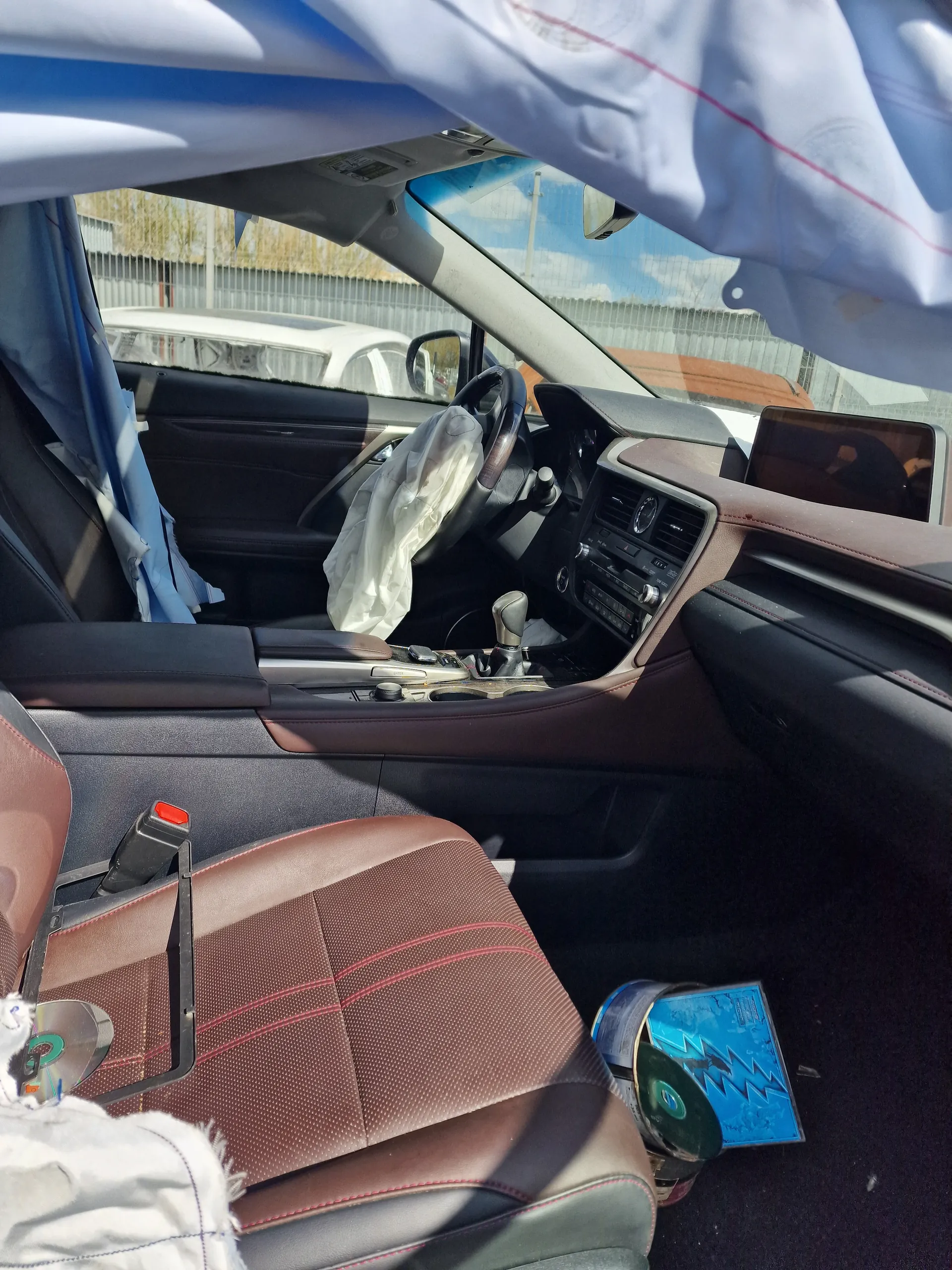 Wnętrze samochodu z brązowymi siedzeniami z czerwonym przeszyciem, kierownicą z rozwiniętą poduszką powietrzną, deską rozdzielczą z ekranem i przyciskami, pudełkiem na dysk CD z niebieskim okładem i błyskawicą oraz dyskiem CD. Widoczne są również fragmenty tkaniny i zewnętrzny widok na budynek przez okno.