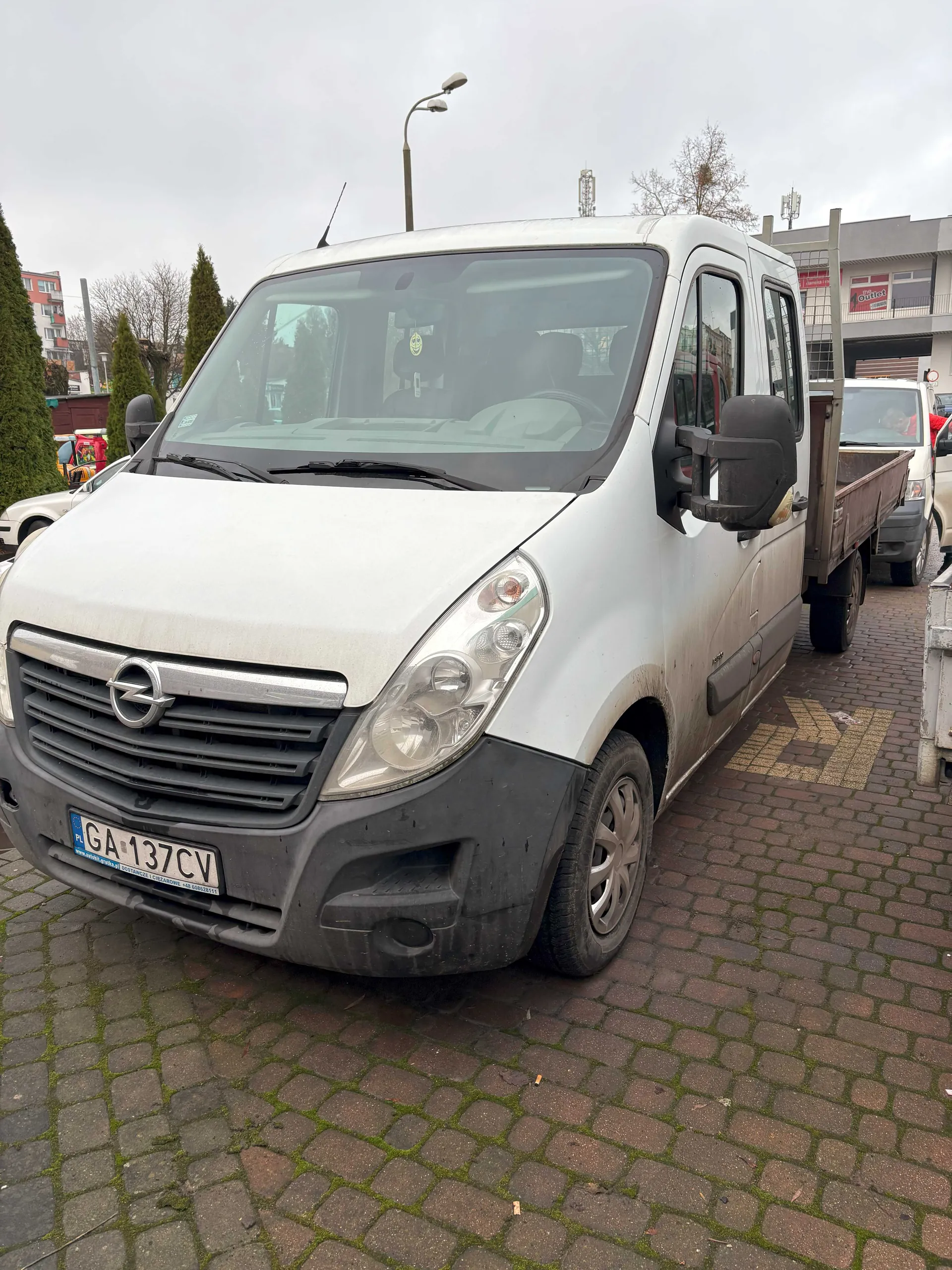 Biały samochód dostawczy marki Opel z tablicą rejestracyjną GA 137CV, zaparkowany na brukowanej ulicy. Widoczne są przednie światła, czarna atrapa chłodnicy z logo Opel, brudne boki pojazdu oraz otwarty tylny zbiornik. W tle budynki, drzewa, latarnia uliczna i inne pojazdy. Niebo jest zachmurzone.