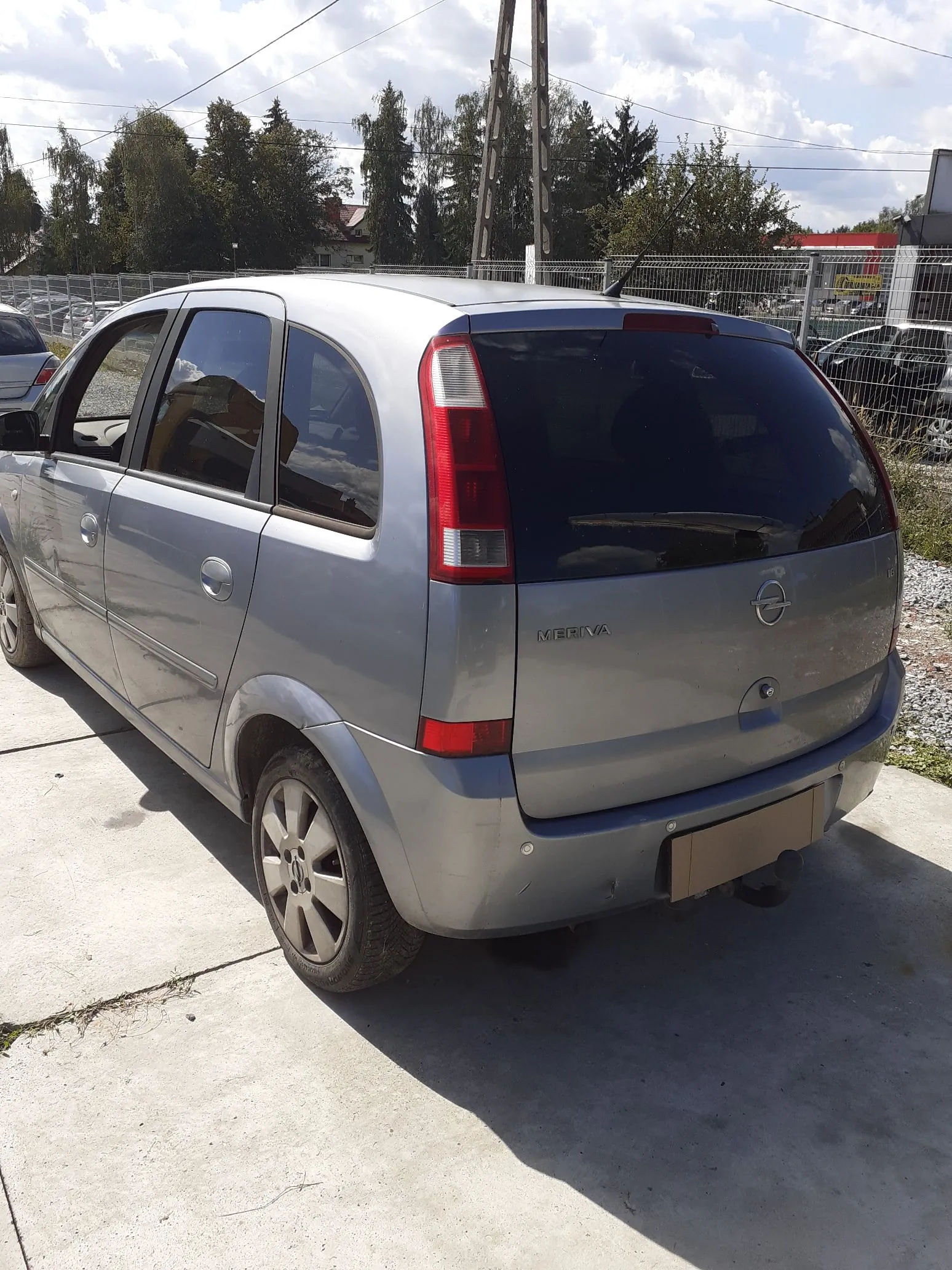 Srebrny samochód marki Opel Meriva zaparkowany na betonowym podłożu. Widok z tyłu i z boku pojazdu. Na klapie bagażnika widnieje napis 'MERIVA' oraz logo producenta. W tle znajduje się ogrodzenie z siatki, drzewa oraz fragmenty innych samochodów pod jasnym, lekko zachmurzonym niebem.