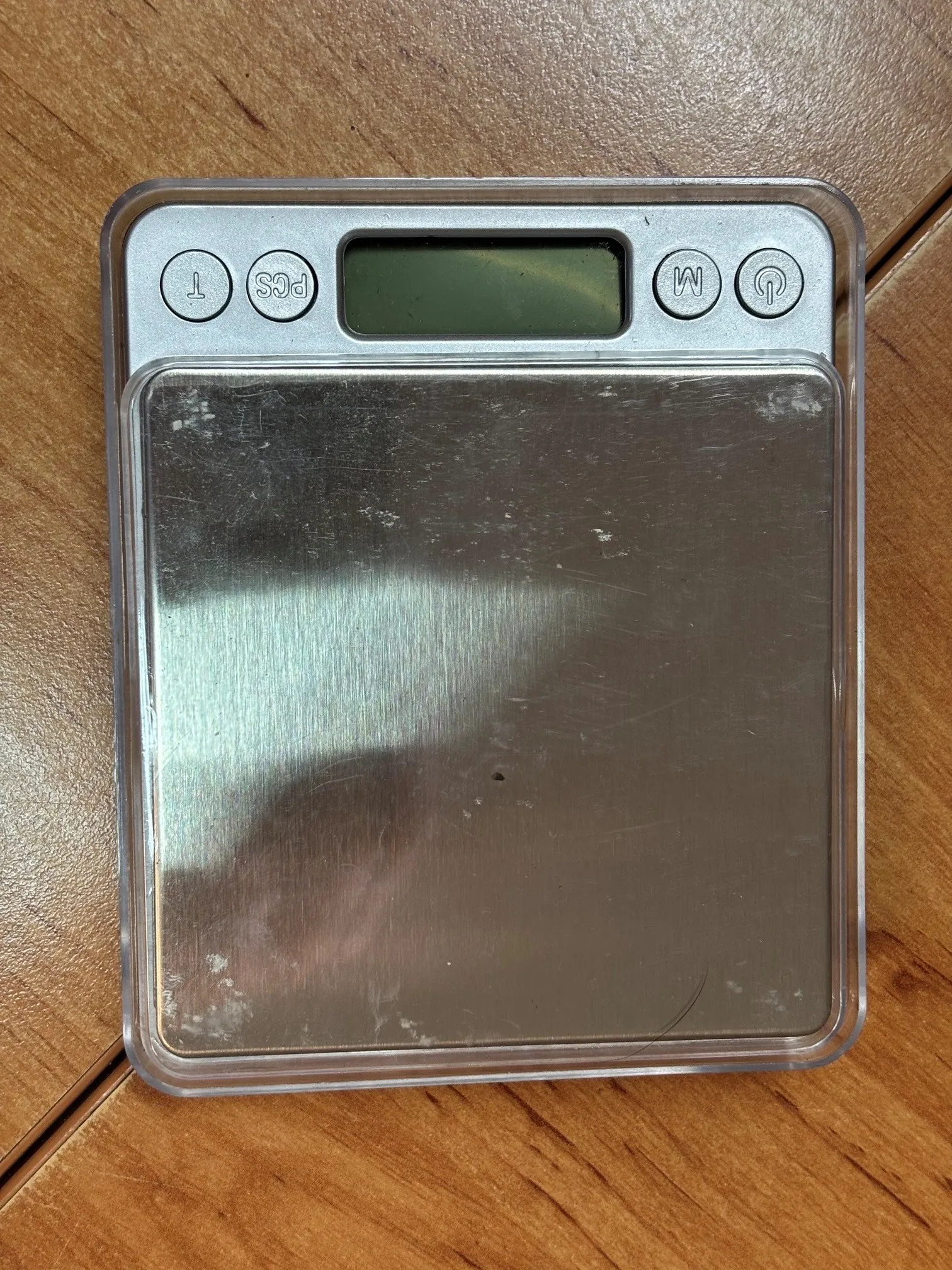 Kwadratowa waga elektroniczna z metalową platformą pomiarową, zamknięta w przezroczystej plastikowej obudowie. Na górze znajduje się ekran LCD oraz cztery okrągłe przyciski z oznaczeniami: 'T', 'PCS', 'M' oraz symbol zasilania.
