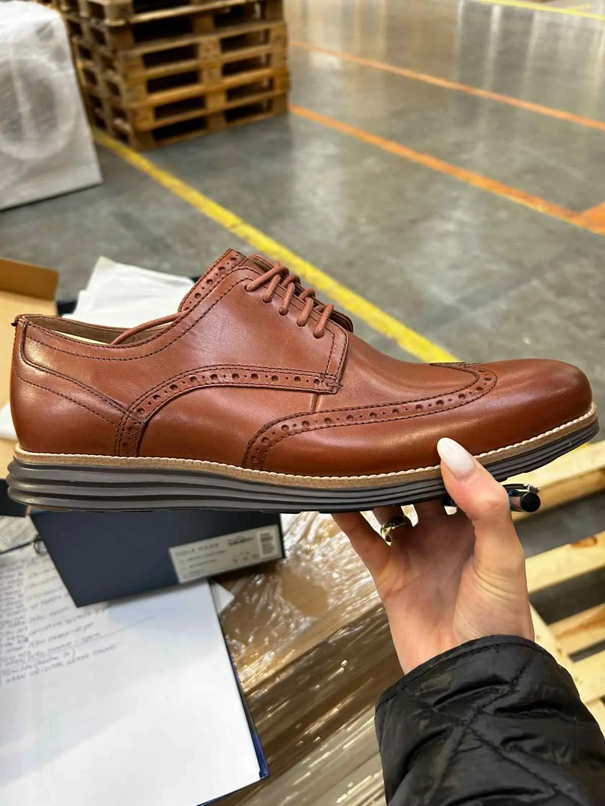 Brązowy skórzany but typu brogue z sznurowaniem, trzymany w dłoni z białym manicurem i pierścieniem. W tle widoczne palety drewniane, betonowa podłoga z żółtymi liniami oraz pudełko z częściowo czytelnym napisem 'GOLF'. Osoba trzymająca but ubrana jest w czarną pikowaną kurtkę.