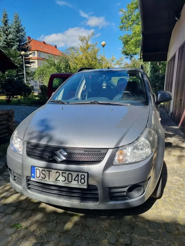 Srebrny samochód marki Suzuki zaparkowany na kostce brukowej. Widoczna jest przednia część pojazdu z polską tablicą rejestracyjną o numerze DST 25048. W tle widać zielone drzewa, fragment czerwonego budynku oraz niebieskie niebo z białymi chmurami.