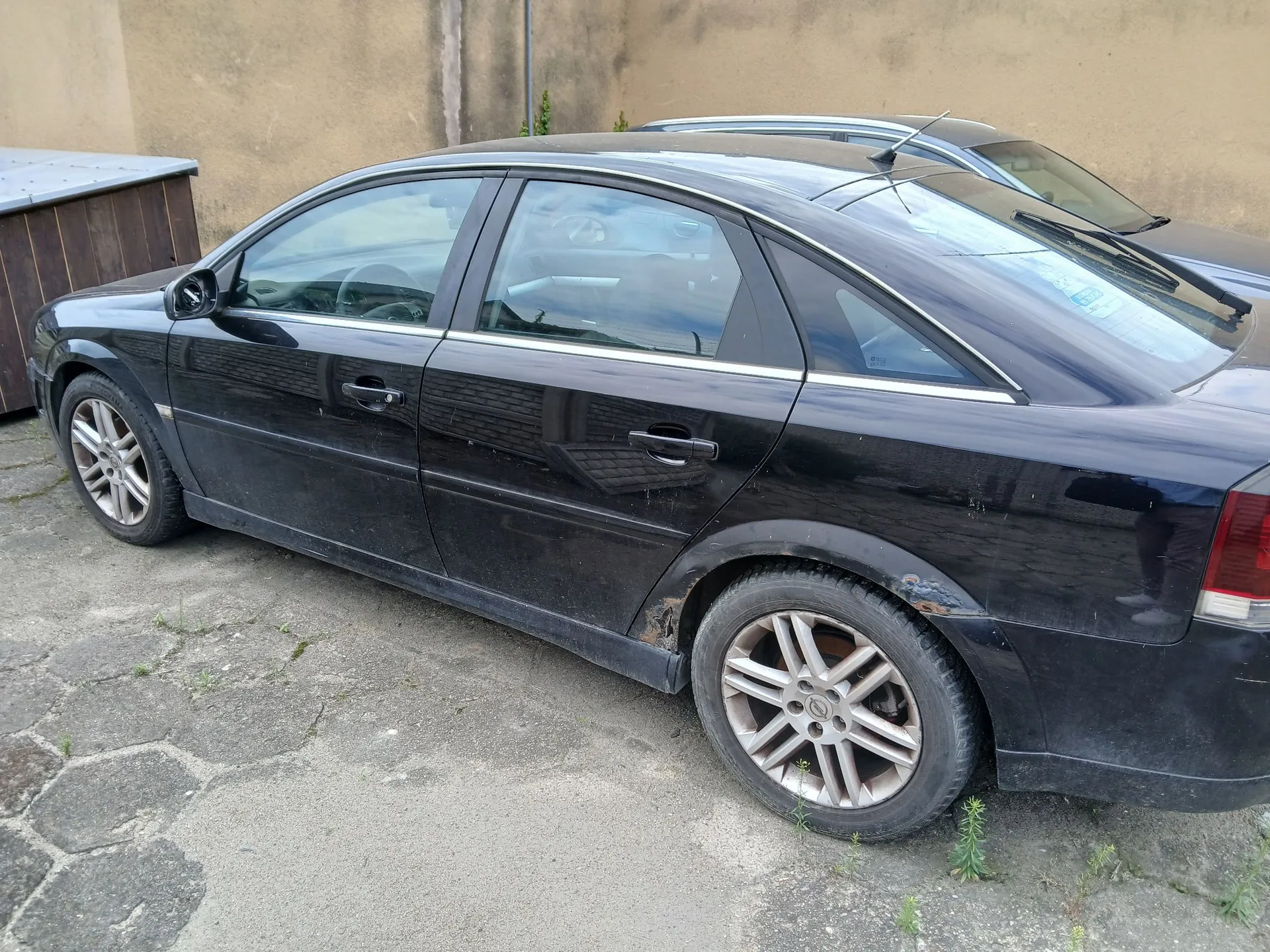 Czarny samochód osobowy typu sedan zaparkowany na betonowym podjeździe. Pojazd jest widoczny z profilu, z tyłu widoczne są ślady korozji nad nadkolem. W tle znajduje się beżowa ściana budynku oraz fragment innego, srebrnego samochodu.
