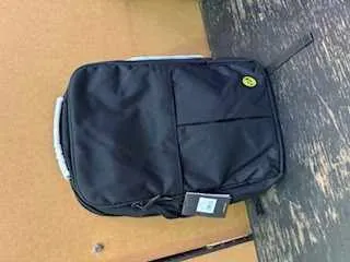 Czarny plecak z przodem kieszenią i żółtym okrągłym logo, umieszczony na drewnianej powierzchni obok brązowego kartonu. Plecak posiada białą etykietę z nieczytelnym tekstem.