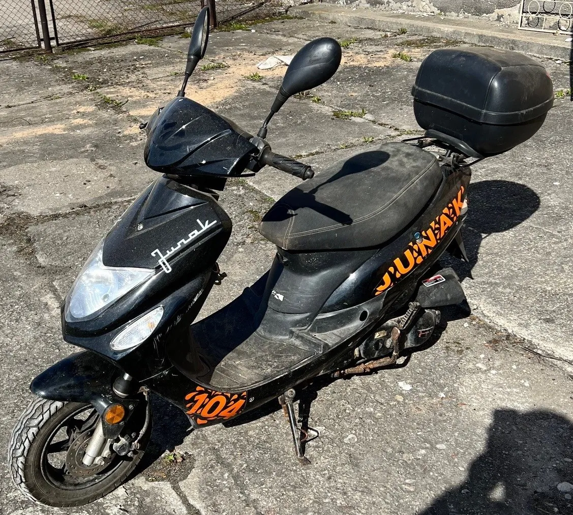Czarny skuter stojący na betonowej powierzchni z widocznymi napisami: 'Juno' na przedniej części, 'JUNAK' oraz '104' na boku w pomarańczowej kolorystyce. Skuter wyposażony w czarny bagażnik na tylnej części, lusterka boczne i czarne siedzenie. W tle betonowa powierzchnia z plamami trawy, metalowe ogrodzenie oraz fragment budynku. Cień skutera i obiektu poza kadrem widoczny na prawo od pojazdu.