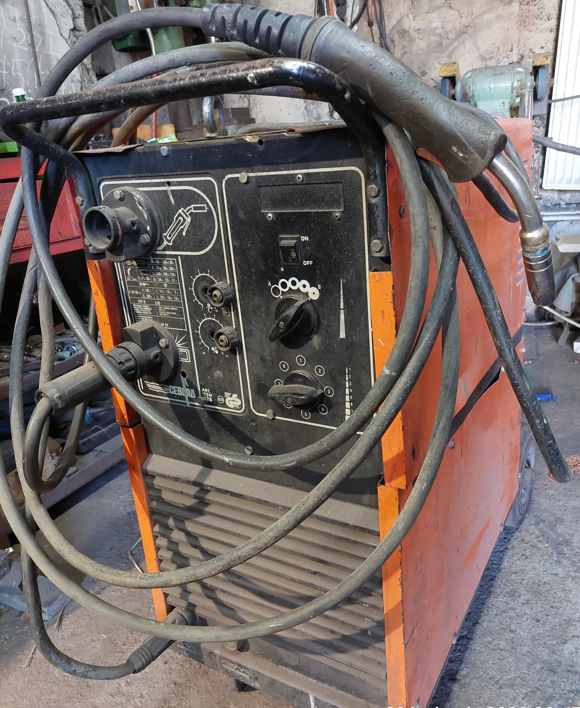 Stacja spawalnicza marki CEBORA o pomarańczowo-czarnym kolorze z przewodami elektrycznymi owiniętymi wokół urządzenia. Na panelu sterowania widoczne są pokrętła, przełącznik ON/OFF, wskaźniki oraz schematyczne oznaczenia. W tle betonowa ściana warsztatu i inne narzędzia. Na panelu widoczny napis CEBORA oraz oznaczenia techniczne.