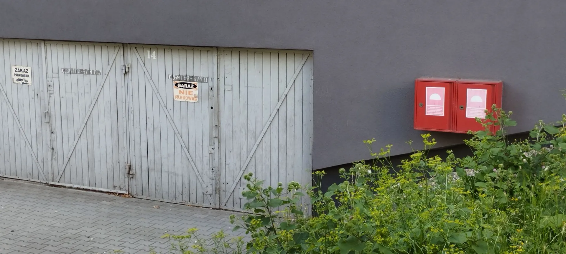 Zdjęcie przedstawia fragment szarej ściany budynku z dużymi, jasnoszarymi drewnianymi drzwiami garażowymi z pionowymi deskami. Na drzwiach znajduje się numer 14 oraz dwie tabliczki z napisami: po lewej „ZAKAZ PARKOWANIA”, a po prawej „GARAŻ NIE PARKOWAĆ”. Po prawej stronie na ścianie zamontowana jest czerwona skrzynka techniczna z dwoma białymi oznaczeniami. Przed budynkiem znajduje się chodnik z kostki brukowej oraz gęste, zielone rośliny z małymi żółtymi kwiatami.