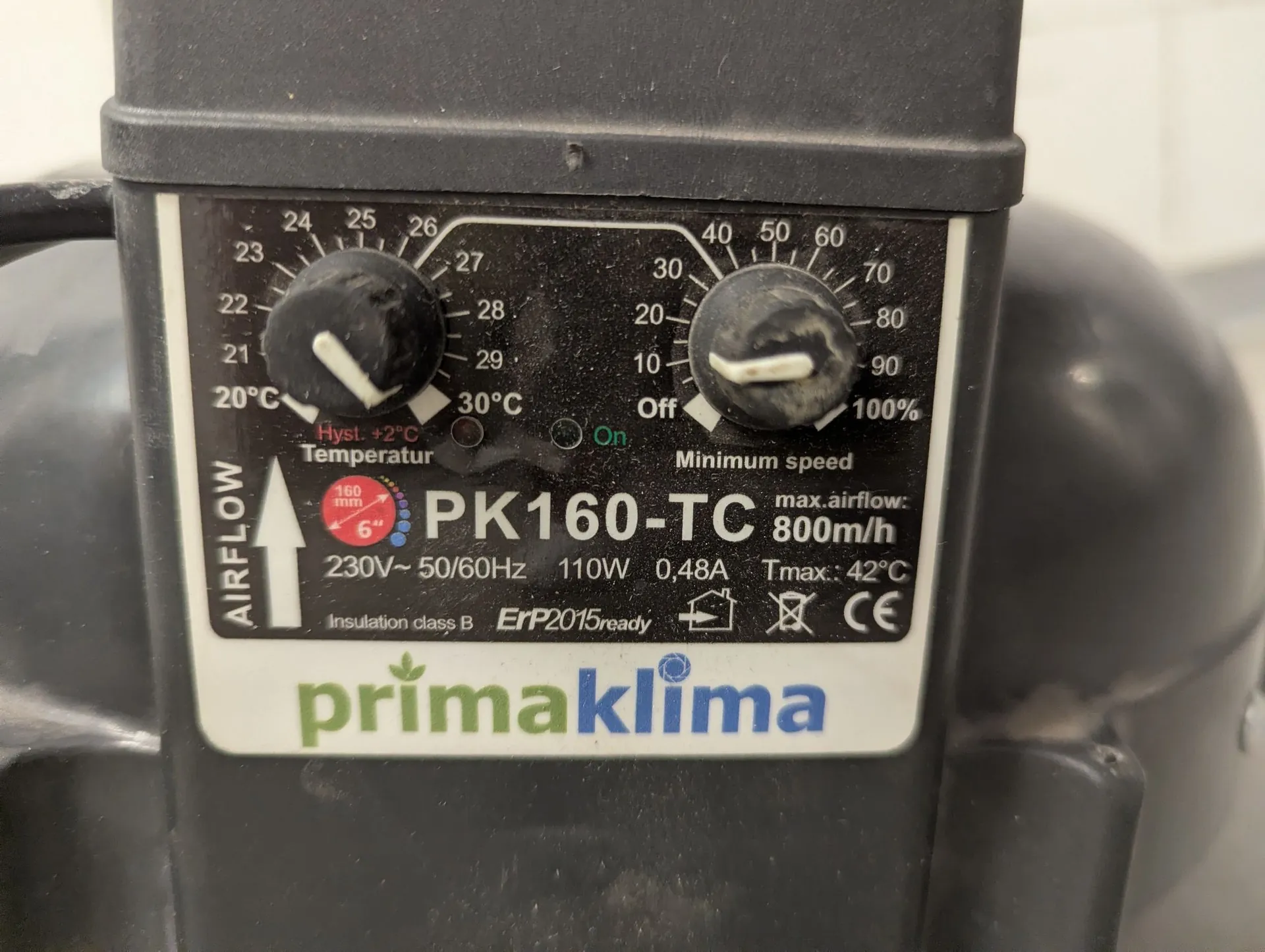 Zbliżenie na panel sterowania urządzenia marki primaklima, model PK160-TC. Panel zawiera dwa pokrętła: lewe do regulacji temperatury w zakresie od 20°C do 30°C oraz prawe do regulacji prędkości od 'Off' do '100%'. Poniżej znajduje się napis 'PK160-TC' oraz dane techniczne: max. airflow 800m/h, zasilanie 230V~ 50/60Hz, moc 110W, natężenie 0,48A oraz temperatura maksymalna Tmax: 42°C. Na dole widnieje duże logo 'primaklima' w kolorach zielonym i niebieskim.
