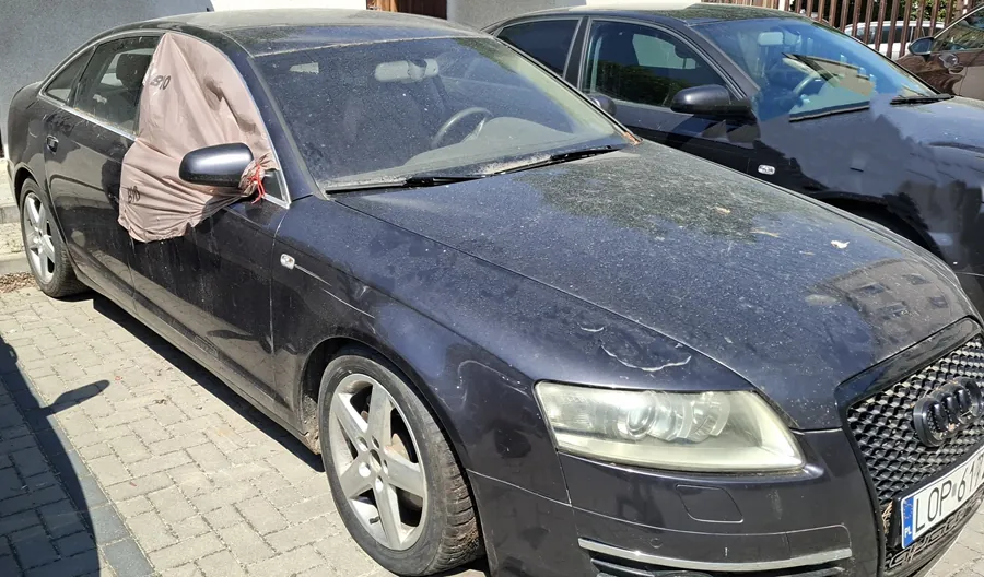 Czarny samochód marki Audi zaparkowany na brukowanej powierzchni. Pojazd pokryty jest kurzem, na lusterku bocznym pasażera znajduje się brązowa tkanina. Widoczna tablica rejestracyjna z napisem LOP 617. W tle drugi czarny samochód, szara ściana budynku oraz drewniane ogrodzenie. Słoneczna pogoda, cienie na ziemi.