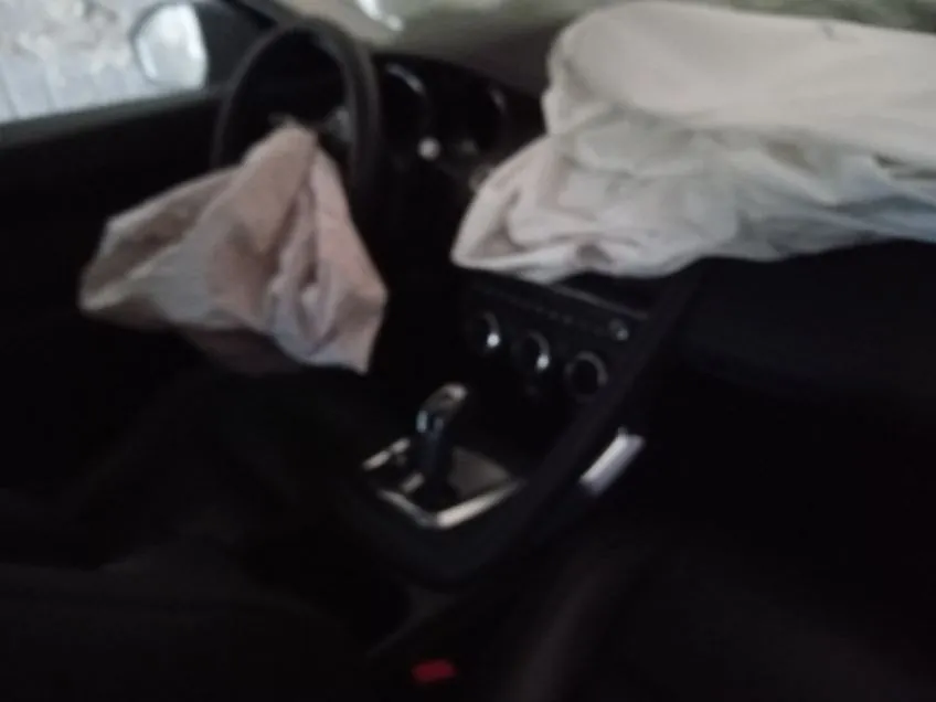 Wnętrze samochodu z rozwiniętymi poduszkami powietrznymi. Widoczne są kierownica po lewej stronie, czarna deska rozdzielcza z przyciskami i dźwignią zmiany biegów. Poduszki powietrzne są rozwinięte: jedna przy kierownicy, druga po stronie pasażera. Czarne siedzenia i fragment czerwonego elementu na siedzeniu. Obraz jest nieco rozmyty.