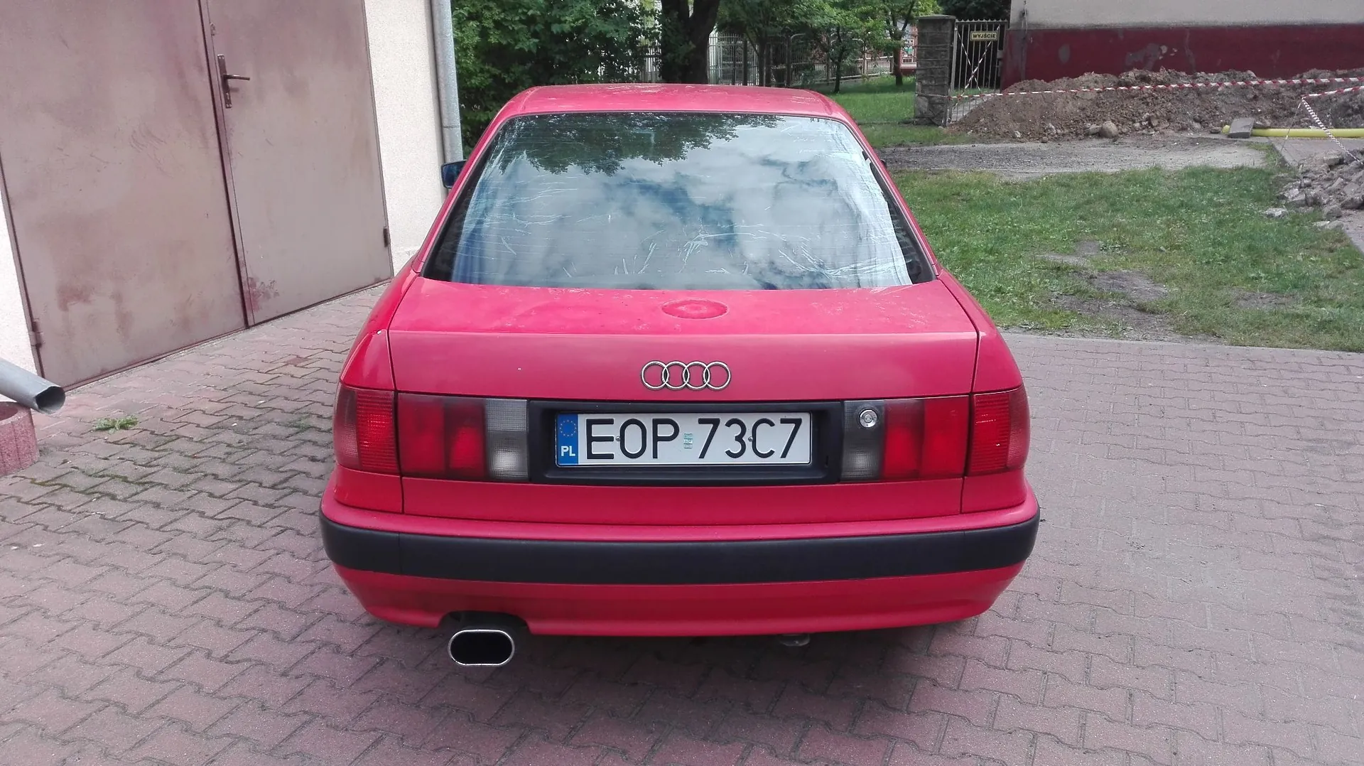 Tylny widok czerwonego samochodu marki Audi zaparkowanego na kostce brukowej. Na klapie bagażnika widoczne jest logo Audi oraz polska tablica rejestracyjna o numerze EOP 73C7. Samochód ma czarny zderzak i jedną rurę wydechową po lewej stronie. W tle widać fragment jasnej ściany budynku, zielone drzewa oraz obszar z ziemią i żwirem.