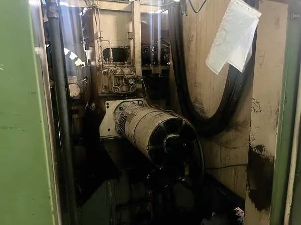 Zdjęcie przedstawiające wnętrze maszyny przemysłowej. Widoczny jest silnik elektryczny z wentylatorem z tyłu, połączony z pompą hydrauliczną. Obok znajdują się przewody, rury oraz fragmenty metalowej obudowy. Na prawej ścianie wisi biała, plastikowa osłona lub filtr.
