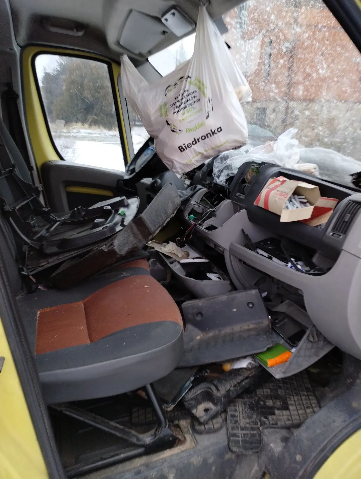 Wnętrze samochodu w stanie dużego nieładu. Na siedzeniu pasażera i podłodze znajdują się liczne części samochodowe, plastikowe elementy i odpady. Z lusterka wstecznego zwisa biała plastikowa torba z logo sieci Biedronka i napisem „TATA... RECYKLING”. Przez przednią szybę widoczny jest zimowy krajobraz z opadami śniegu.