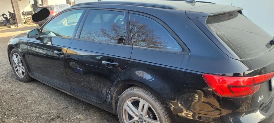 Czarny samochód Audi A4 Avant (kombi) widziany z boku i tyłu, zaparkowany na podwórku z żwiru. Widoczne są odbicia drzew w szybach, inny samochód oraz budynek z cegły na tle. Na tylnej części pojazdu widnieje napis 'A4'.