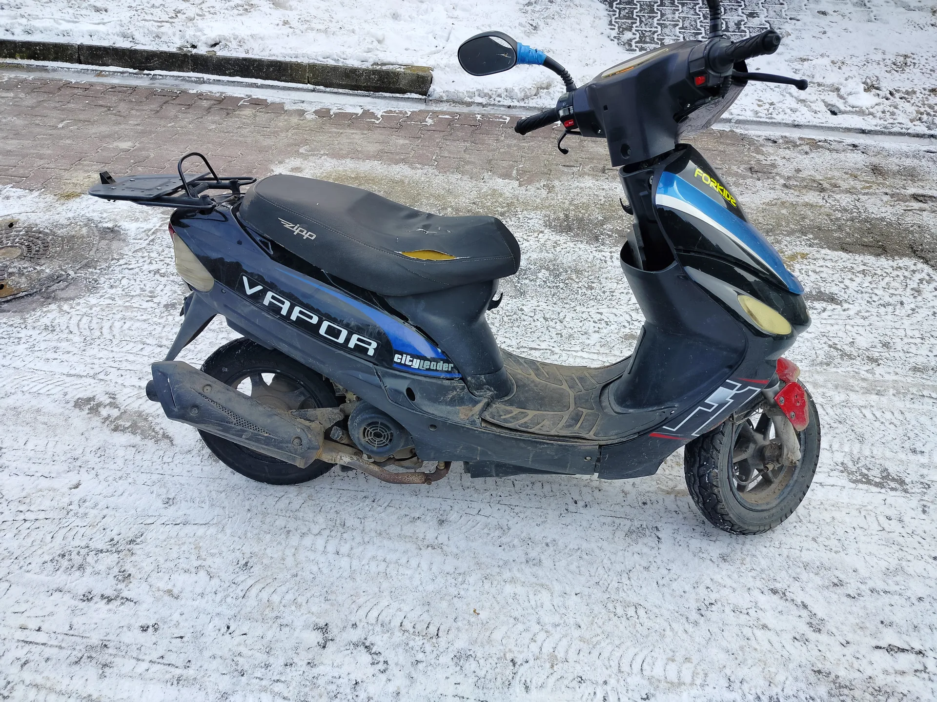 Czarny skuter z niebieskimi akcentami marki VAPOR, z widocznym napisem cityleader na boku i ZIPP na siedzeniu. Na przedniej części skutera widoczny żółty napis FOR. Siedzenie ma uszkodzenie z odsłoniętym żółtym materiałem. Skuter stoi na śnieżnej nawierzchni z widocznymi odciskami opon, w tle brukowana droga i śnieżny chodnik. Na skuterze widoczne lusterko, uchwyt na bagaż oraz czerwone elementy na przednim kole.