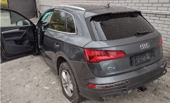 Szary samochód marki Audi Q5 zaparkowany na piaszczystym podłożu obok jasnej ściany z cegieł. Przednie lewe drzwi samochodu są otwarte. Widoczny jest tył pojazdu z logo Audi, oznaczeniem modelu Q5 oraz pustą ramką na tablicę rejestracyjną.