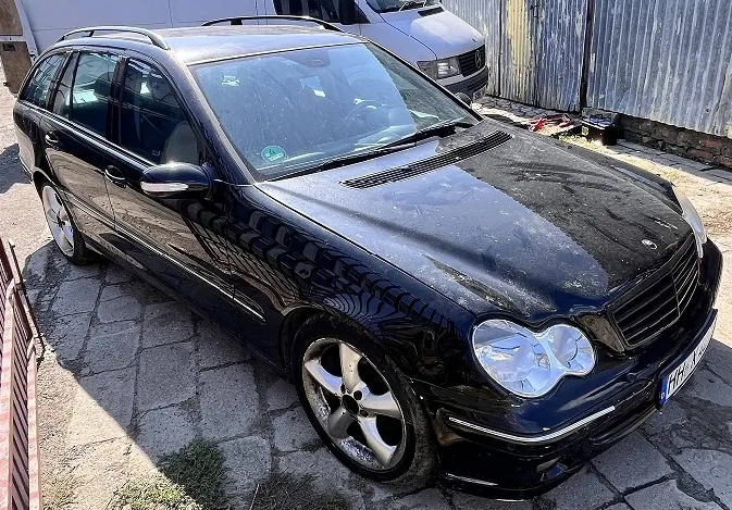 Czarny samochód marki Mercedes-Benz w nadwoziu kombi, zaparkowany na kostce brukowej. Pojazd jest zakurzony, widoczne są srebrne felgi aluminiowe oraz fragment tablicy rejestracyjnej. W tle znajduje się biały samochód dostawczy oraz metalowy płot.