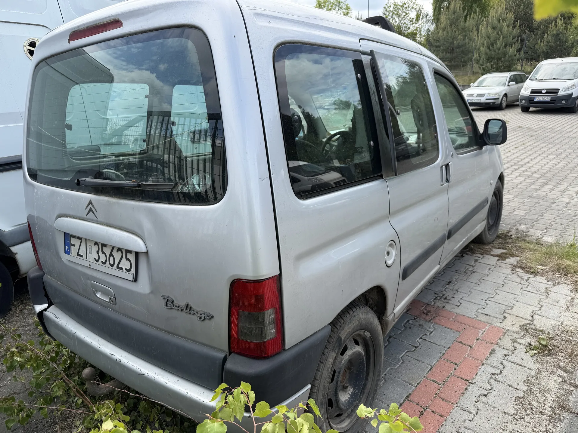 Srebrny samochód dostawczy Citroën Berlingo zaparkowany na brukowanej powierzchni. Widoczny tył pojazdu z tablicą rejestracyjną PL FZI 35625, czerwonymi światłami tylnymi oraz napisem "Berlingo" na bagażniku. W tle inne pojazdy, drzewa i częściowo zachmurzone niebo. Na pierwszym planie zielone liście roślin.