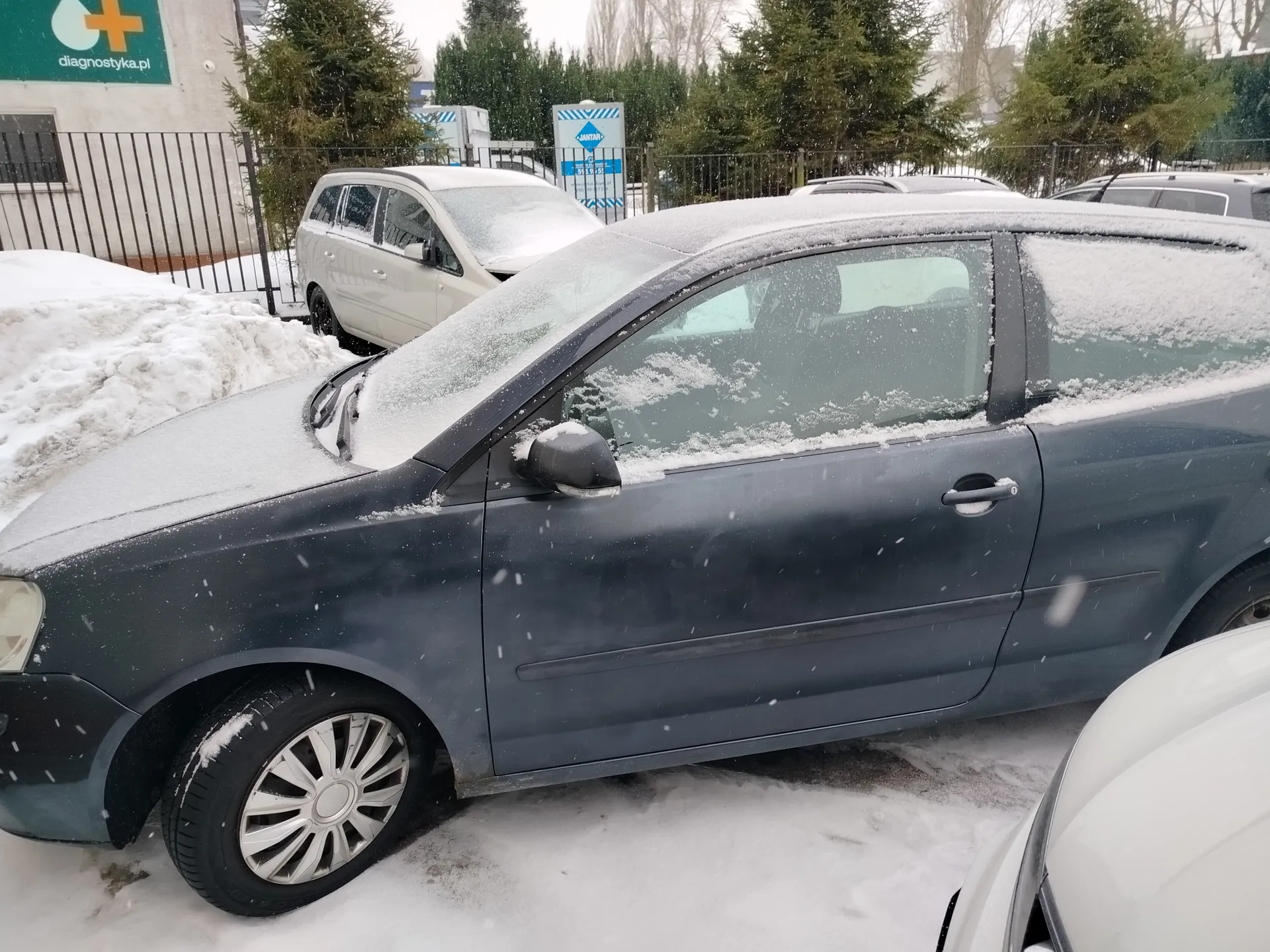 Zdjęcie przedstawia parking pokryty śniegiem z kilkoma zaparkowanymi samochodami. Na pierwszym planie znajduje się ciemnoszary samochód osobowy z warstwą śniegu na dachu i szybach. W tle widoczny jest biały samochód, ogrodzenie metalowe, zielony znak z napisem 'diagnostyka.pl' oraz niebieski znak z napisem 'JANTAR'. Wokół rosną drzewa iglaste, a na ziemi leży śnieg. Pada śnieg.