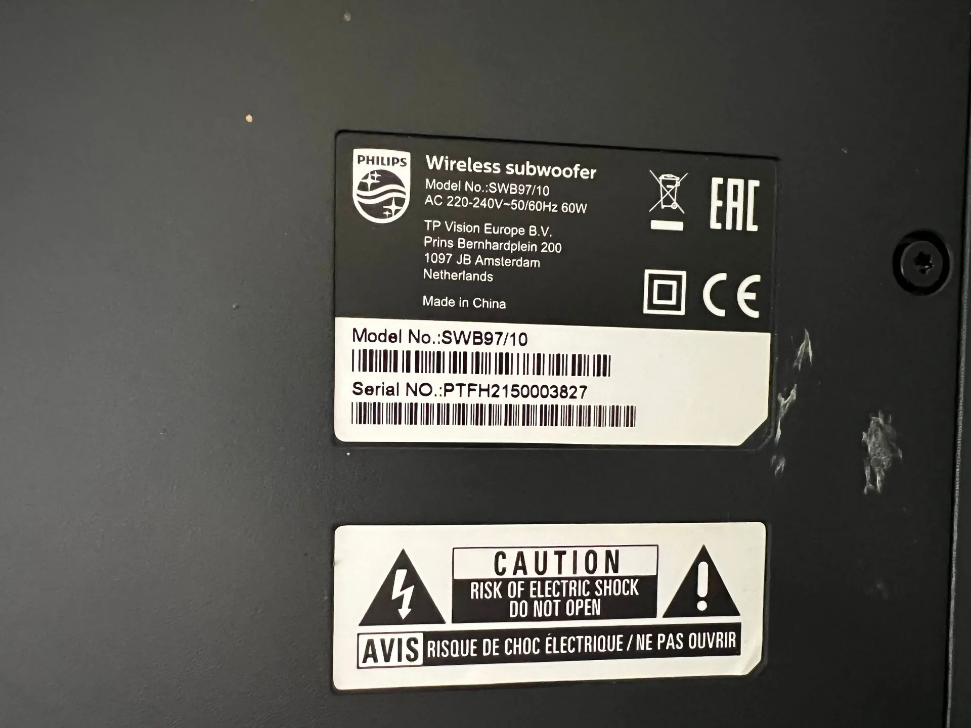 Zdjęcie tylnej części czarnego urządzenia elektronicznego z dwiema naklejkami informacyjnymi. Górna naklejka zawiera logo Philips oraz tekst: 'Wireless subwoofer', 'Model No.: SWB97/10', 'AC 220-240V~50/60Hz 60W', 'TP Vision Europe B.V., Prins Bernhardplein 200, 1097 JB Amsterdam, Netherlands', 'Made in China'. Poniżej znajduje się numer modelu 'Model No.: SWB97/10' oraz numer seryjny 'Serial NO.: PTFH2150003827' wraz z kodami kreskowymi. Po prawej stronie widoczne są symbole EAC i CE. Dolna naklejka to ostrzeżenie z ikonami błyskawicy w trójkątach i tekstem: 'CAUTION RISK OF ELECTRIC SHOCK DO NOT OPEN' oraz poniżej w języku francuskim: 'AVIS RISQUE DE CHOC ÉLECTRIQUE / NE PAS OUVRIR'.
