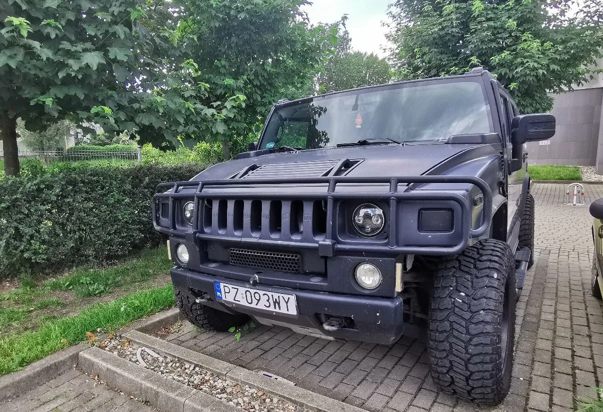 Czarny samochód terenowy marki Hummer zaparkowany na kostce brukowej. Pojazd posiada masywny przedni zderzak z rurą ochronną, charakterystyczny grill z pionowymi szczelinami oraz duże, terenowe opony. Na zderzaku znajduje się polska tablica rejestracyjna o numerze PZ 093WY. W tle widoczne są zielone drzewa i krzewy.