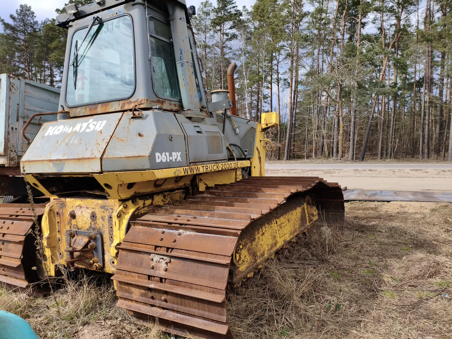 Stary, zardzewiały buldożer gąsienicowy marki Komatsu model D61-PX o kolorze szaro-żółtym. Na maszynie widoczne napisy: 'KOMATSU', 'D61-PX' oraz 'WWW.TRUCK-2000.DE'. Gąsienice i część przednia maszyny pokryte są rdzą. W tle las z wysokimi drzewami, droga oraz sucha trawa na ziemi. Na szybie kabiny widoczny numer '3314'.