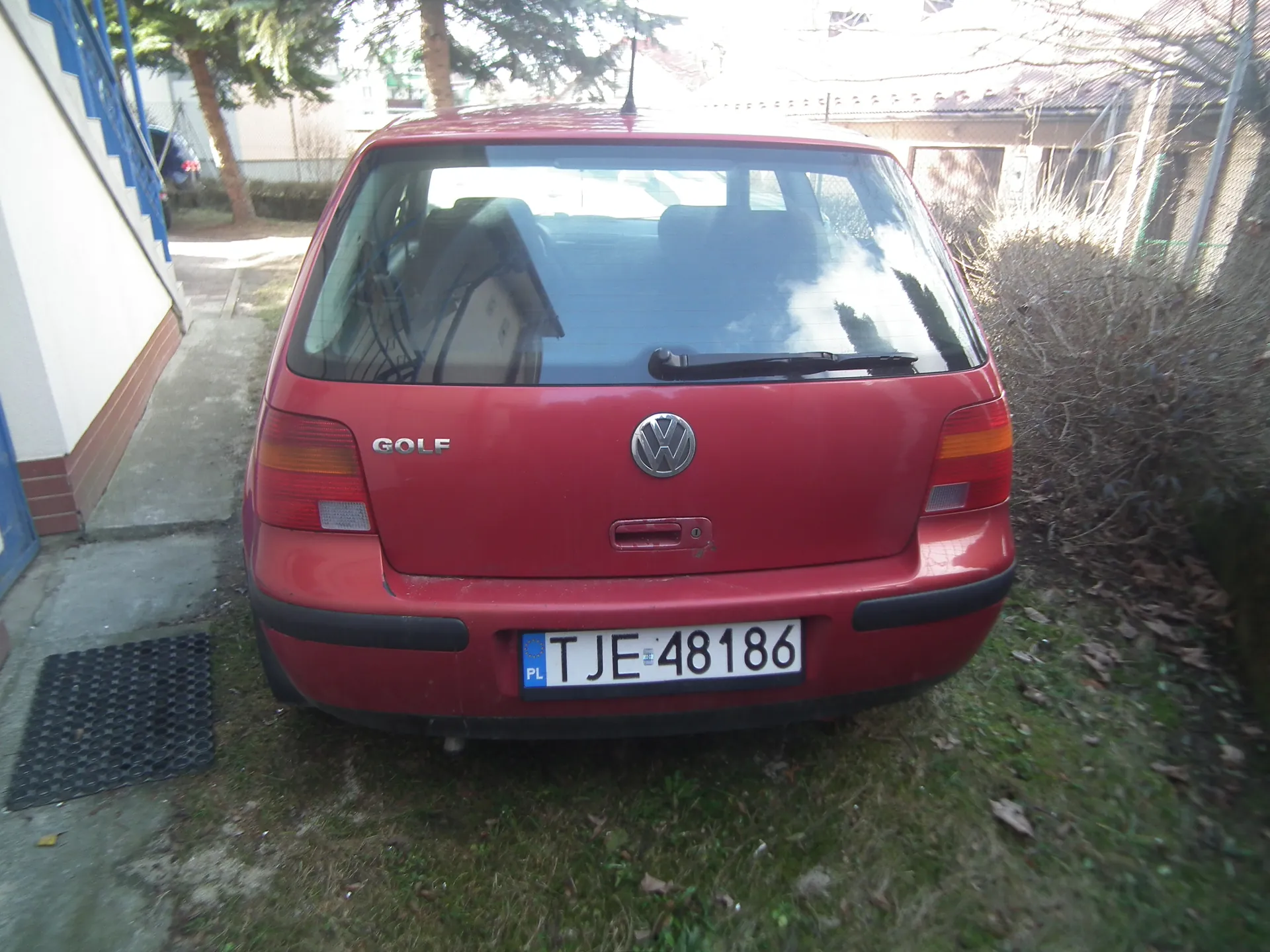 Czerwony samochód Volkswagen Golf zaparkowany na trawie, widok od tyłu. Na bagażniku widoczne logo VW i napis GOLF. Tablica rejestracyjna z polskim kodem PL oraz numerem TJE 48186. Po lewej stronie budynku z niebieskimi schodami, w tle drzewa, krzewy i domy. Słoneczny dzień, na trawie rozrzucone suche liście.