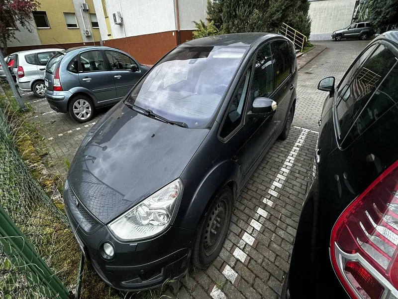 Czarny minivan zaparkowany na brukowanym placu parkingowym, otoczony innymi samochodami (szary hatchback, srebrny samochód). W tle budynek z żółto-białą elewacją, zieleń, ogrodzenie i żółte schody. Na pierwszym planie fragment czarnego samochodu z czerwonym światłem tylnym. Parkingu towarzyszą białe linie wyznaczające miejsca postojowe.