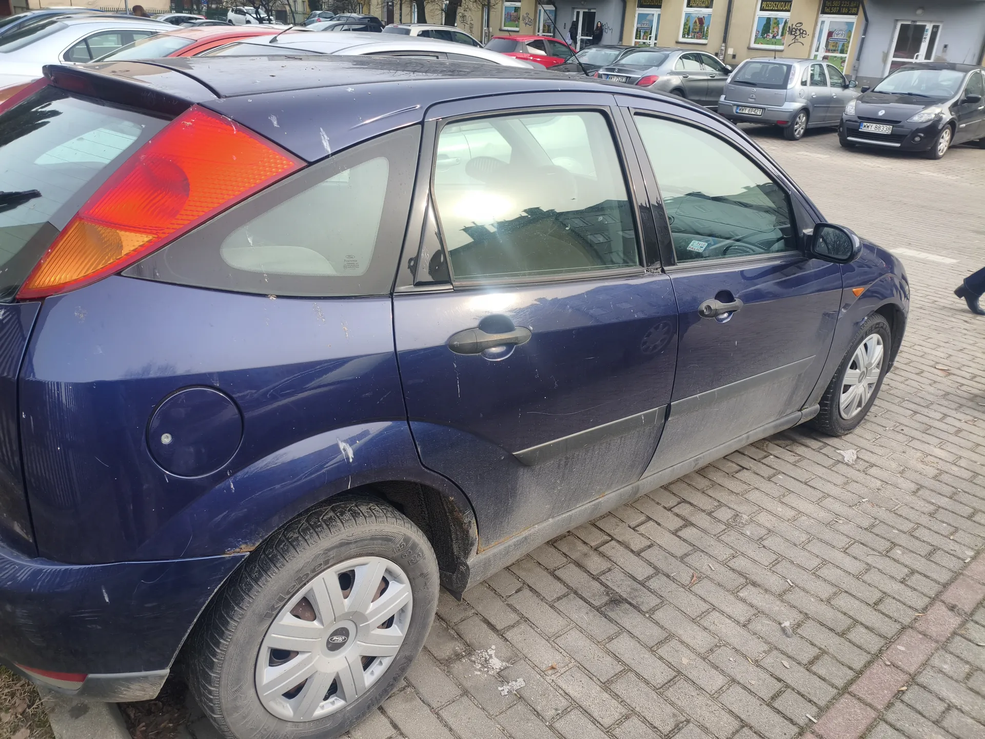Niebieski samochód osobowy marki Ford, model Focus, zaparkowany na brukowanej powierzchni. Samochód ma zabrudzone dolne partie nadwozia i opony. Widoczne są tylne światło z czerwonym i pomarańczowym segmentem, srebrne felgi oraz pokrywa zbiornika paliwa. W tle stoją inne pojazdy, w tym czarny samochód z tablicą rejestracyjną WY 8838 oraz budynki z oknami i szyldami sklepowymi. Na prawym brzegu kadru widoczna jest część nogi przechodnia.