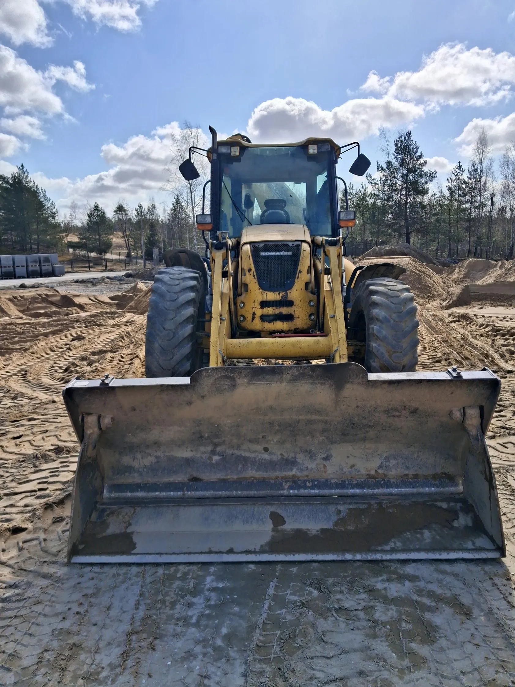 żółty pojazd budowlany z dużą metalową łopatą na przodzie, stojący na piaskowatej powierzchni z odciskami opon. W tle widoczne są stosy piasku, drzewa, niebo z białymi chmurami oraz budynki po lewej stronie