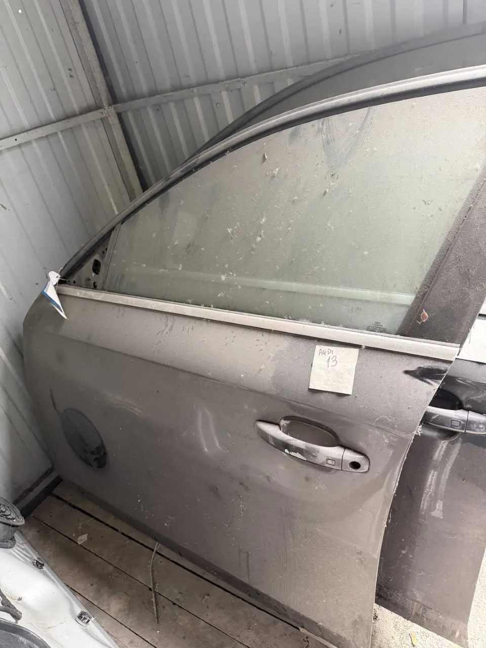 Szara drzwi samochodowe z uchwytem i szybą, oparte o metalową ścianę z blachy falistej. Na drzwiach naklejka z napisem 'A4 13'. W tle widoczne drewniane deski podłogi oraz fragment innego pojazdu. Drzwi pokryte są kurzem i drobnymi uszkodzeniami.
