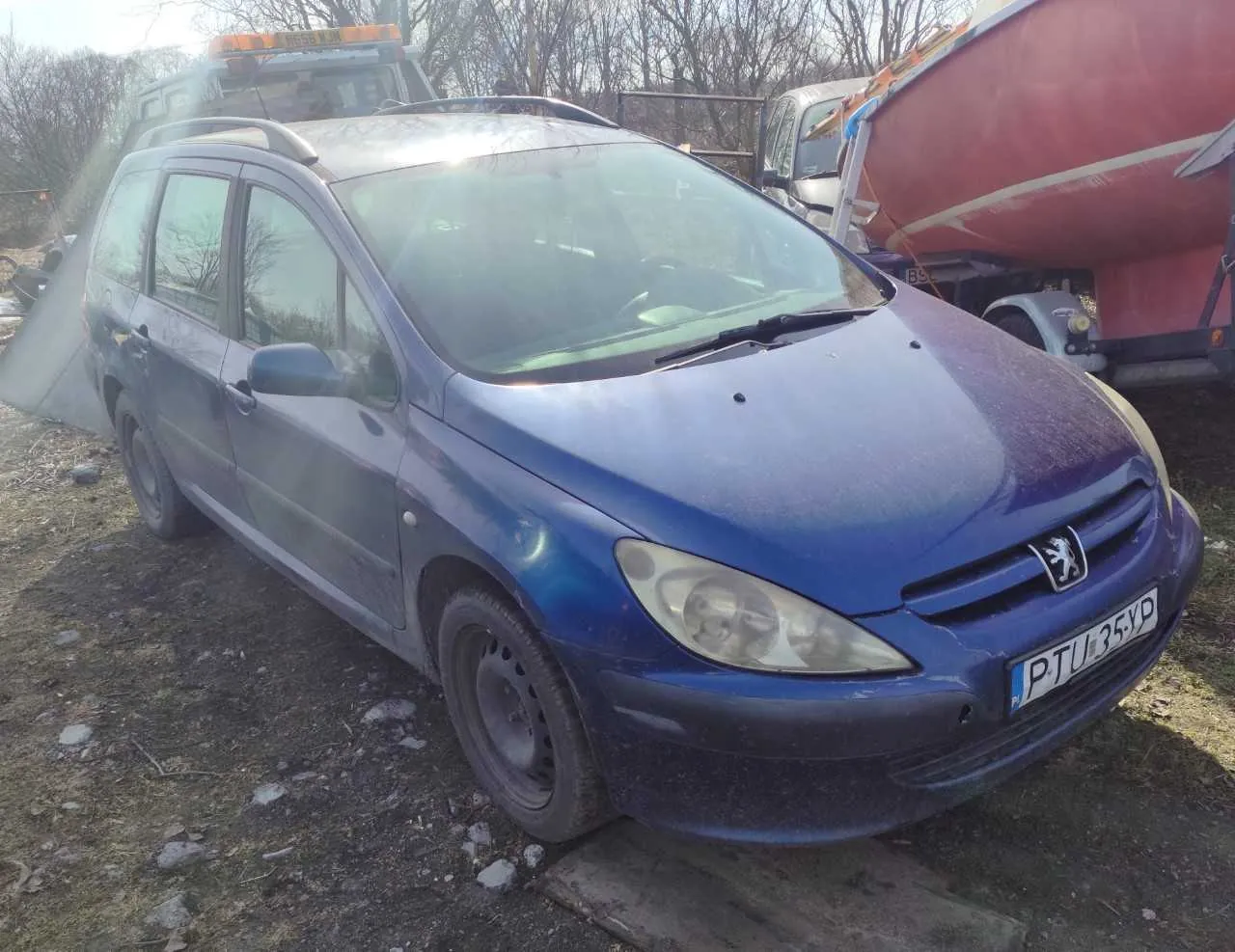 Niebieski samochód osobowy marki Peugeot, typ kombi, z widocznym logo na masce i tablicą rejestracyjną P-TU 35YD. Pojazd znajduje się na nierównym terenie z kamieniami i suchą trawą, przednim kołem opartym na drewnianej desce. W tle widoczna czerwona łódź na przyczepie, inne pojazdy oraz drzewa bez liści. Słońce świeci, co powoduje odblask na przedniej szybie.