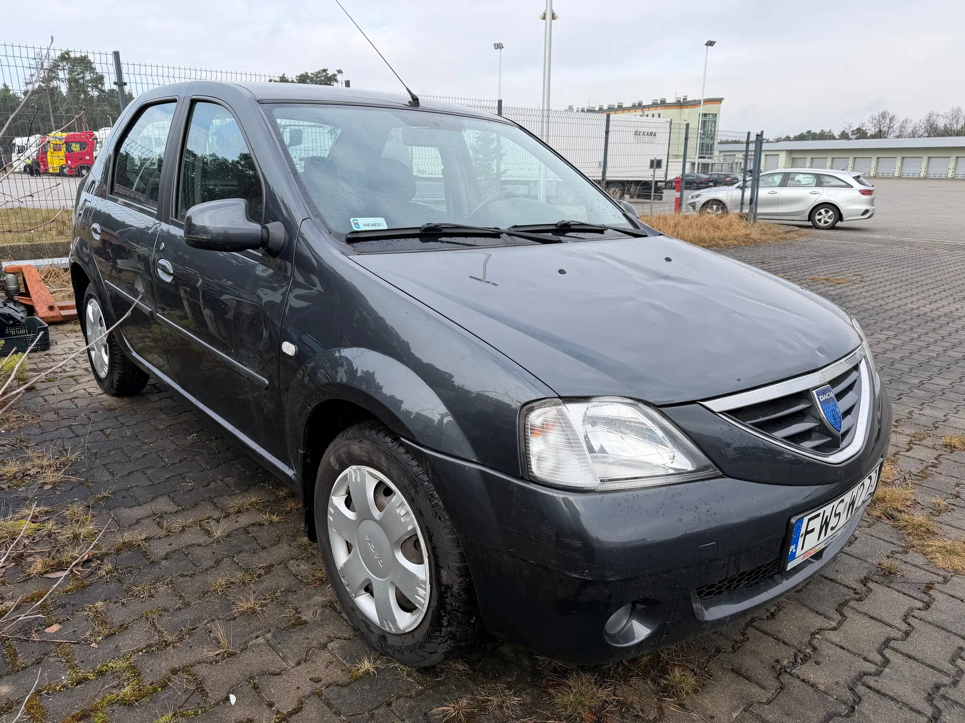 Ciemnoszary samochód marki Dacia z tablicą rejestracyjną FWS W22, zaparkowany na brukowanej powierzchni z widocznymi plamami trawy. W tle ogrodzenie siatkowe, srebrny samochód, czerwono-żółty pojazd ciężarowy oraz biały naczepowy z napisem OZ KARA. Widoczne również przemysłowe budynki i słupy oświetleniowe pod pochmurnym niebem.
