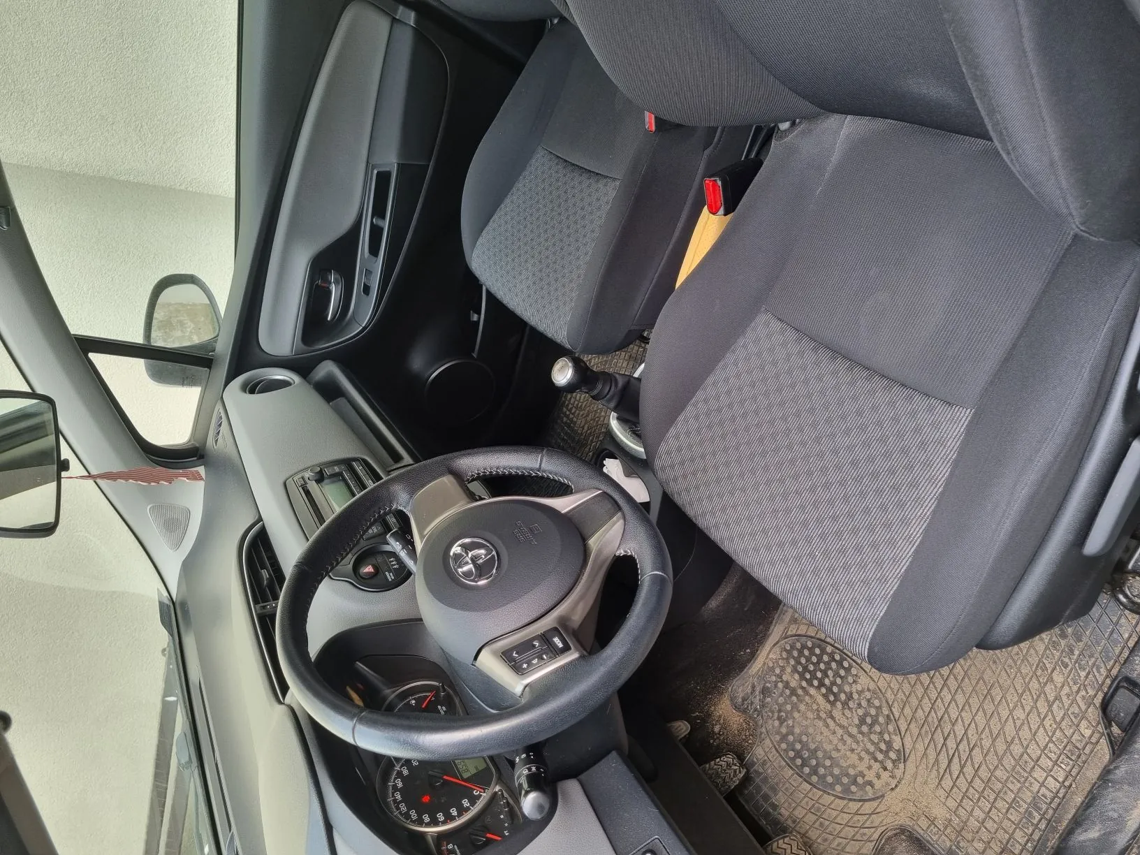 Wnętrze samochodu z widocznym kierownicą z logo Toyoty, czarnymi siedzeniami z szarym materiałem, deską rozdzielczą z licznikami, przyciskami i radioodtwarzaczem. Widoczne są również otwarte drzwi pasażera, lusterko boczne, podłoga z brudnymi dywanikami oraz czerwone zapięcia pasów bezpieczeństwa. W tle przez otwarte drzwi widać fragment ściany budynku.