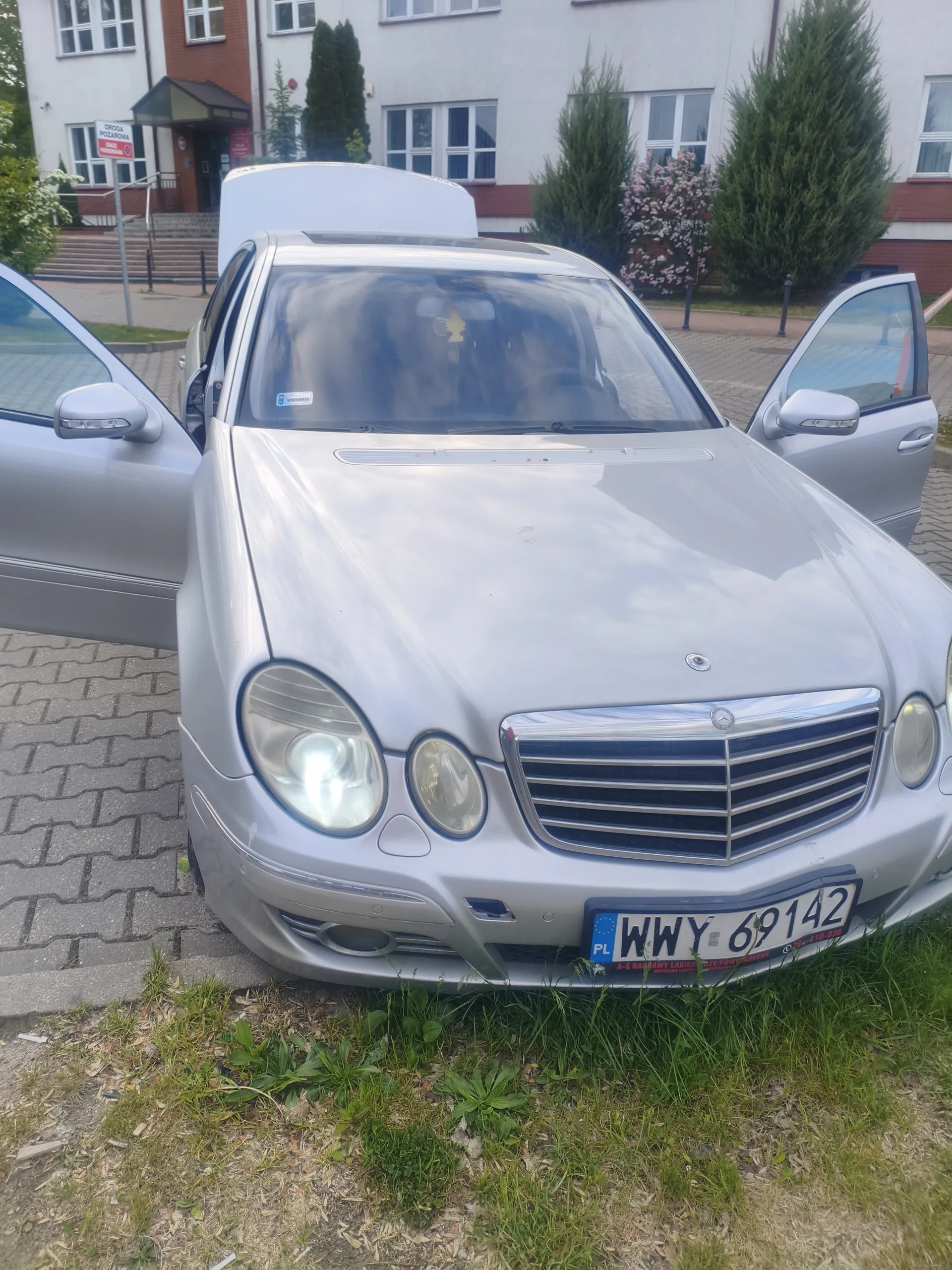Srebrny samochód marki Mercedes-Benz z otwartymi drzwiami przodowymi i bagażnikiem, zaparkowany na brukowanej powierzchni. Widoczna tablica rejestracyjna PL WWY 69142. W tle budynek z oknami, drzewa, krzewy oraz znak drogowy z napisem DROGA POŻAROWA. Przed samochodem trawa i ziemia.