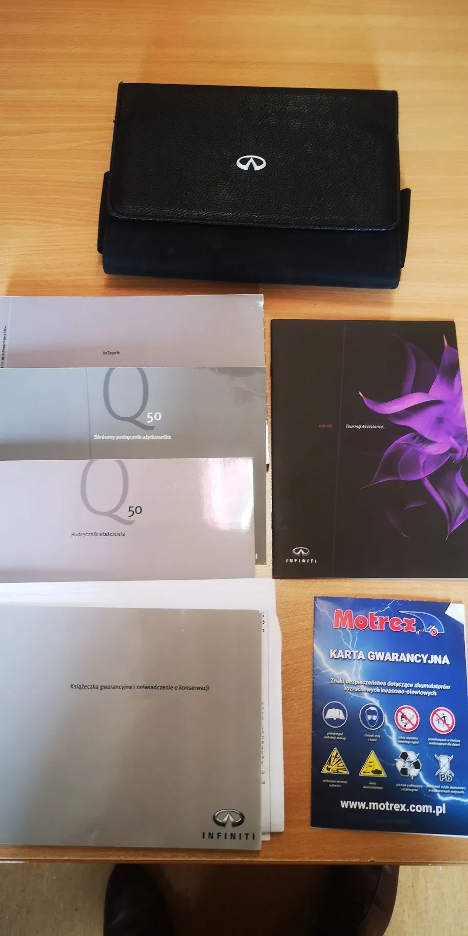 Zestaw dokumentów i akcesoriów marki Infiniti ułożonych na drewnianym blacie. Na górze znajduje się czarne, skórzane etui z logo Infiniti. Poniżej, po lewej stronie, widnieją trzy broszury: jedna z napisem 'inTouch', druga z napisem 'Q50 Skrócony podręcznik użytkownika' oraz trzecia z napisem 'Q50 Podręcznik właściciela'. Po prawej stronie znajduje się ciemna folder z fioletowym kwiatem i napisem 'Touring Assistance' oraz logo Infiniti. Na samym dole po lewej stronie leży 'Książeczka gwarancyjna i zaświadczenie o konserwacji' z logo Infiniti, a obok niej niebieska karta gwarancyjna firmy Motrex z napisem 'KARTA GWARANCYJNA' i adresem strony internetowej 'www.motrex.com.pl'.