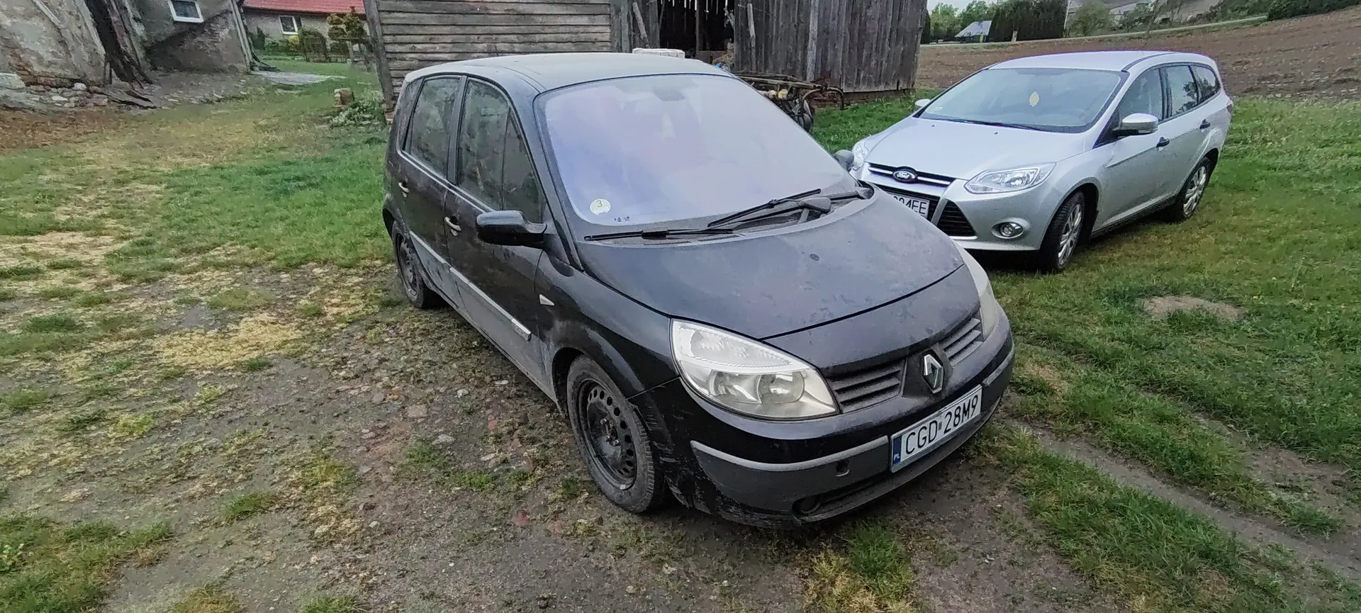 Dwa samochody zaparkowane na trawiastym terenie z widocznymi plamami ziemi. Przedni, czarny samochód marki Renault z tablicą rejestracyjną CGD 28M9, z lekkim zabrudzeniem nadwozia i naklejką na przedniej szybie. Za nim srebrny samochód marki Ford z częściowo widoczną tablicą 304EE. W tle drewniana stodoła, fragmenty budynków i zielona trawa. Horyzont jest poziomy, tekst na tablicach czytelny.
