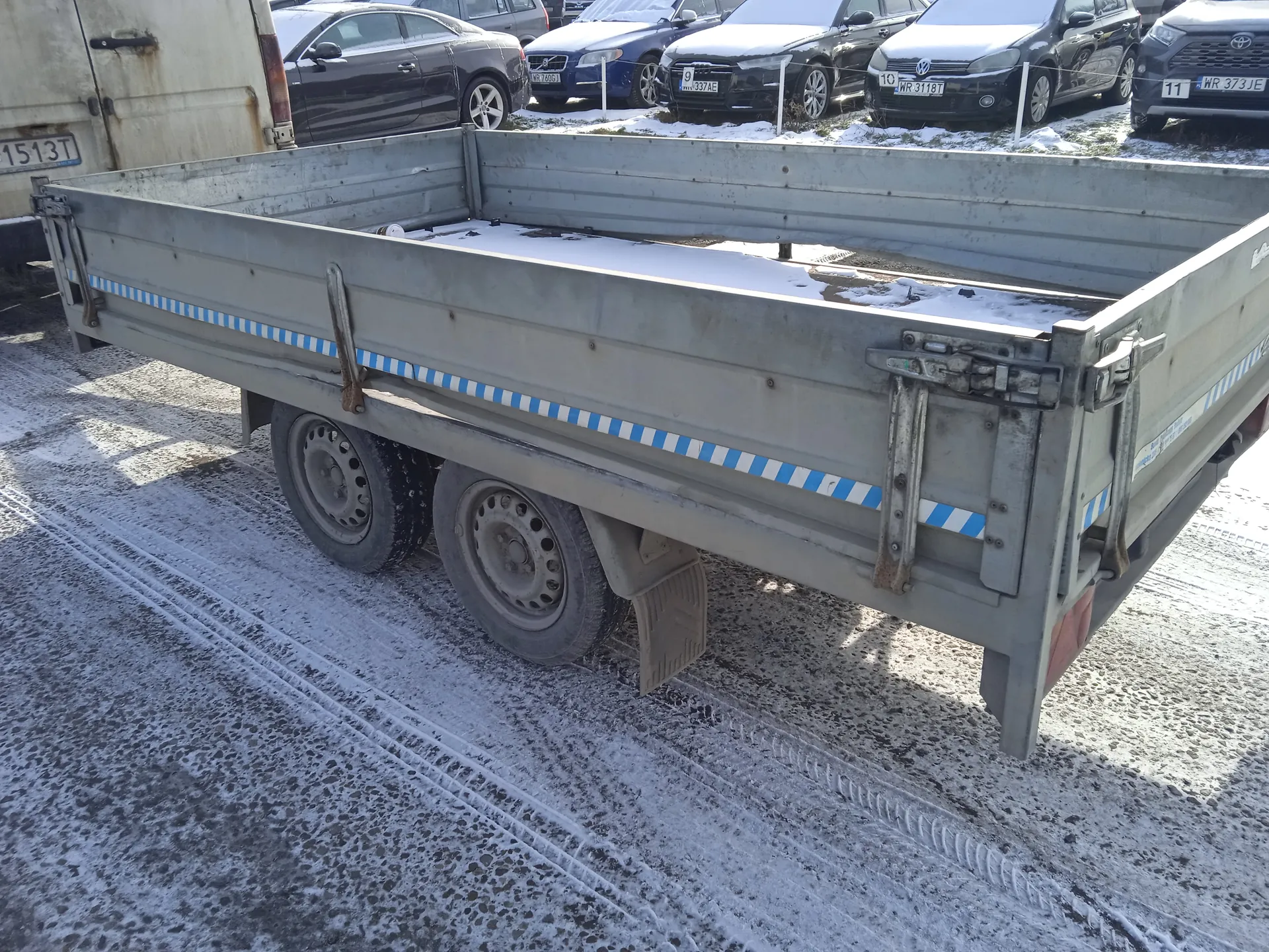 Szary metalowy przyczepa z niebiesko-białymi pionowymi paskami, stojąca na śnieżnej nawierzchni z widocznymi śladami opon. W tle zaparkowane samochody z tablicami rejestracyjnymi: WR 3118T, WR 373 JE, 1513T oraz inne. Na przyczepie i ziemi leży śnieg. Przyczepa ma dwa koła i metalowe zapięcia po bokach.
