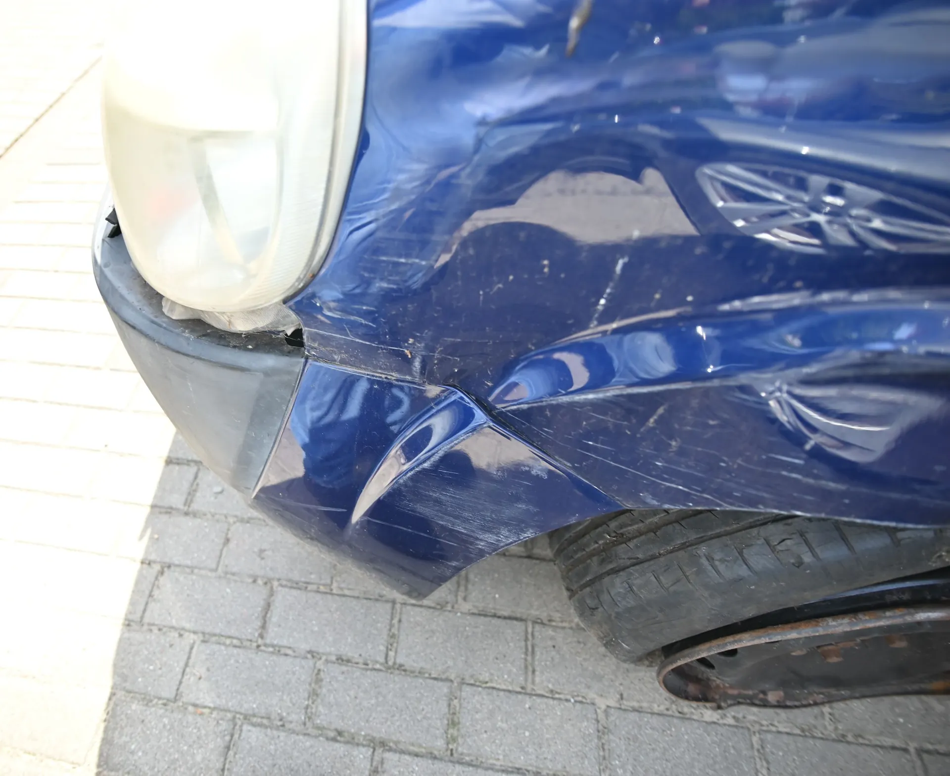 Zbliżenie na uszkodzony przód niebieskiego samochodu. Widoczne jest wgniecenie i zarysowania na błotniku oraz zderzaku, a także pęknięcie w miejscu styku reflektora z karoserią. Samochód stoi na szarej kostce brukowej.