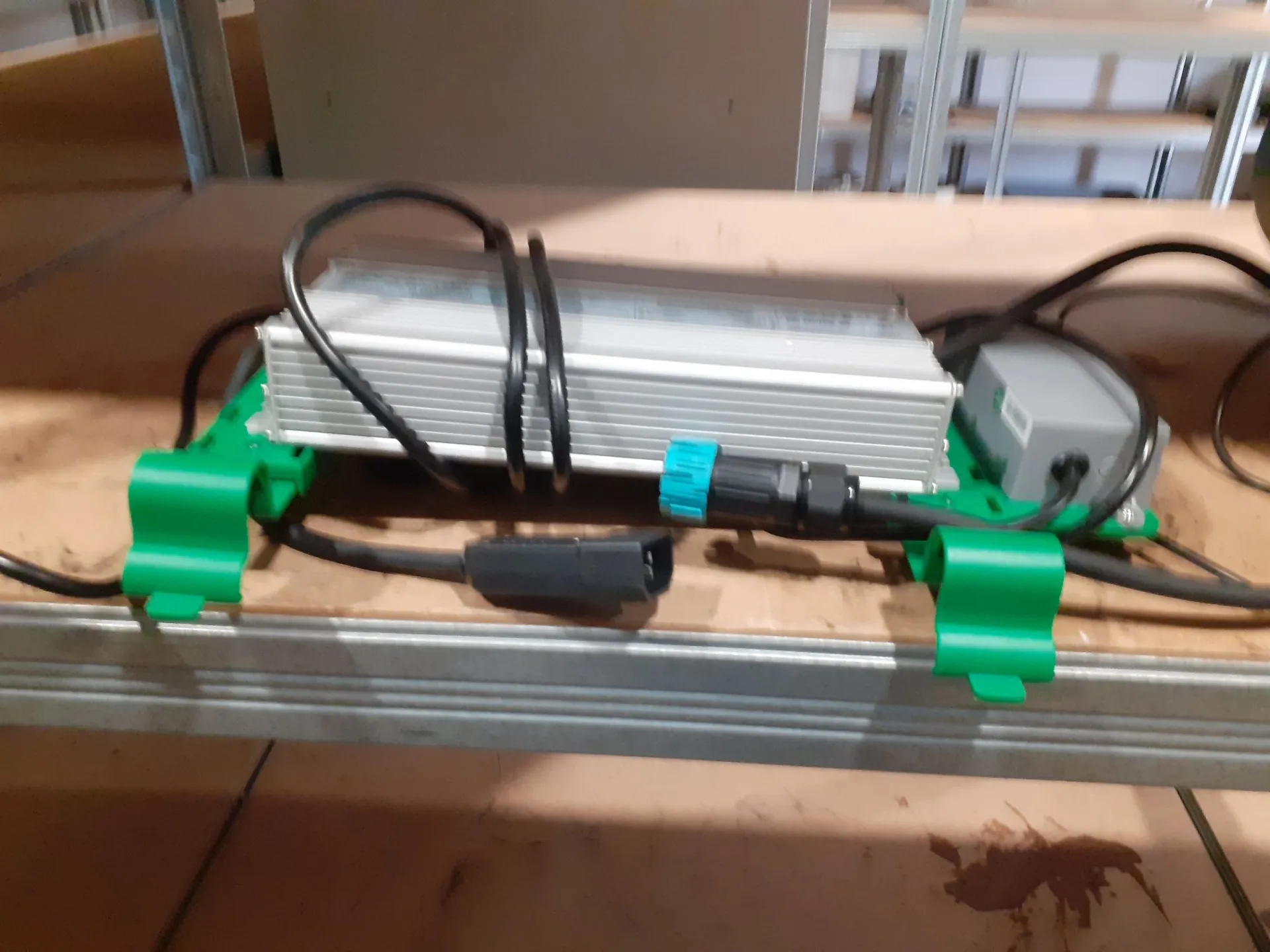 Urządzenie elektroniczne w aluminiowej obudowie z radiatorami, zamocowane do metalowej szyny za pomocą dwóch zielonych uchwytów plastikowych. Do urządzenia podłączone są czarne kable, w tym jeden z niebieskim elementem złączki. Całość spoczywa na brązowej powierzchni.