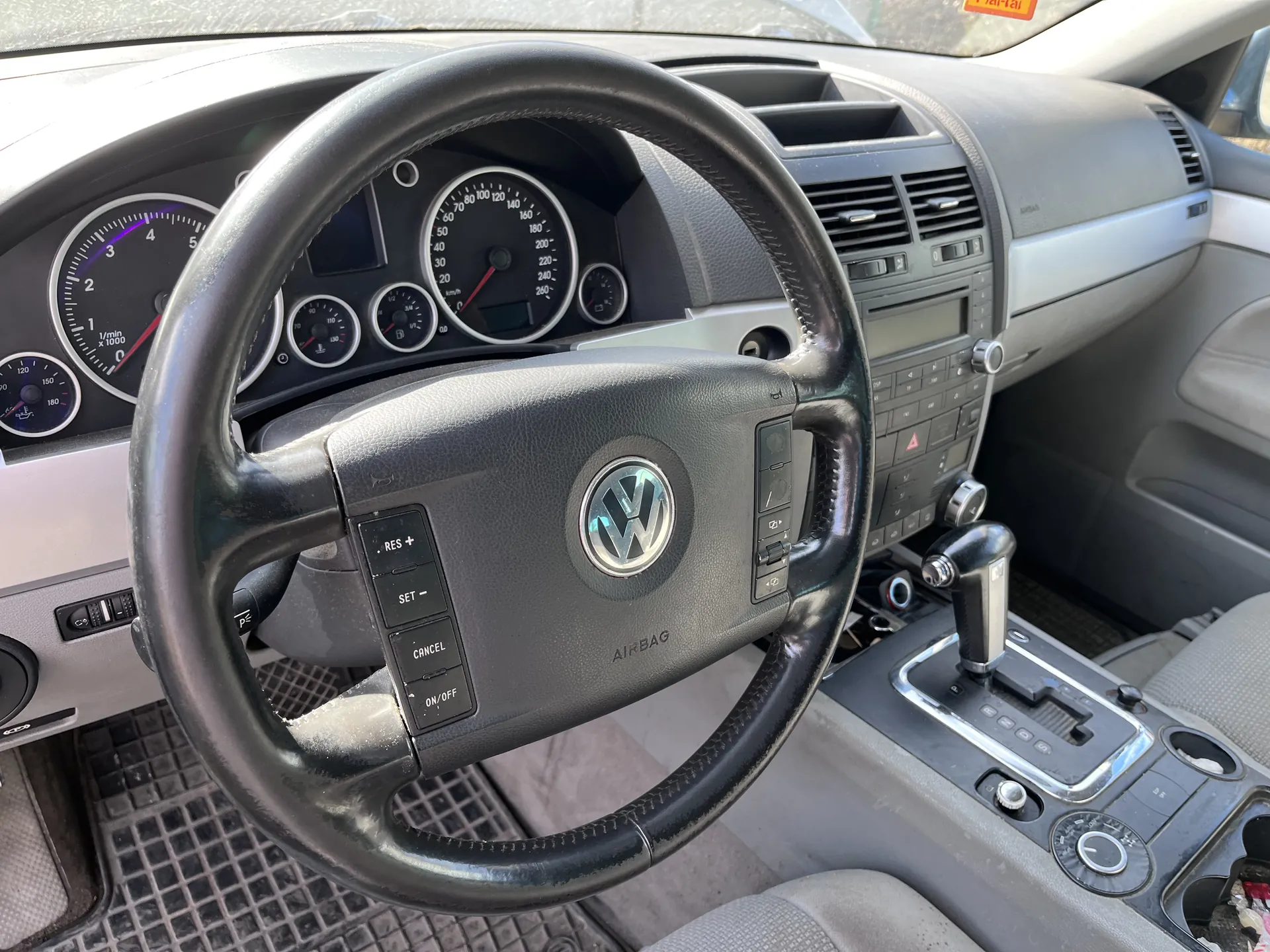 Wnętrze samochodu marki Volkswagen z widocznym kierownicą z logo VW, przyciskami RES+, SET-, CANCEL, ON/OFF oraz napisem AIRBAG. W tle zegary samochodowe: prędkościomierz, obrotomierz i inne wskaźniki. Widoczna również deska rozdzielcza z klimatyzacją, radio i dźwignią zmiany biegów. Na przedniej szybie znajduje się pomarańczowy naklejony znaczek.