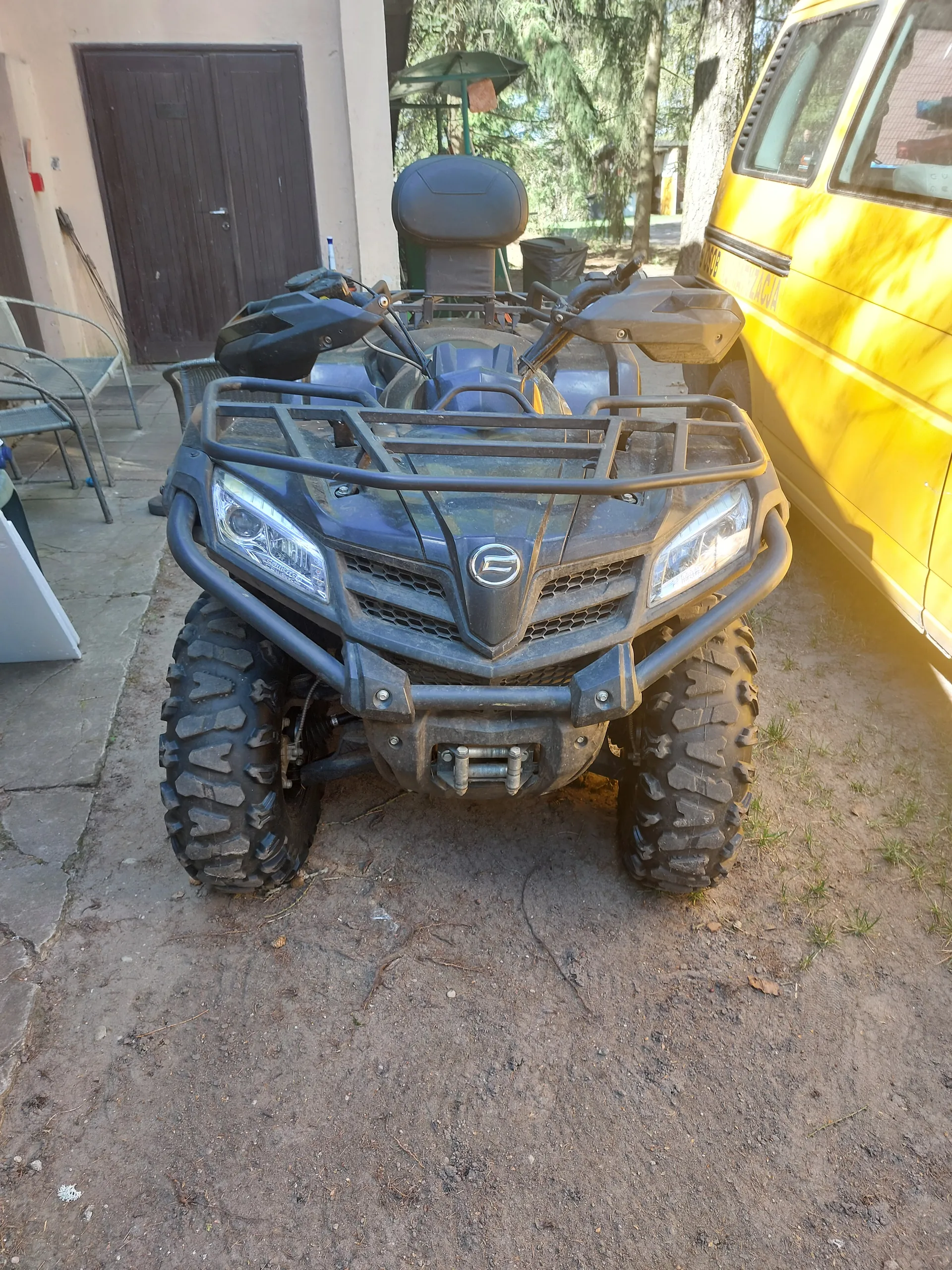 Czarny quad z dużymi oponami terenowymi, z przodu widoczna osłona i reflektory, zaparkowany na brudnej ziemi obok częściowo widocznego żółtego pojazdu. W tle budynek z brązowymi drzwiami, drzewa i inne przedmioty.