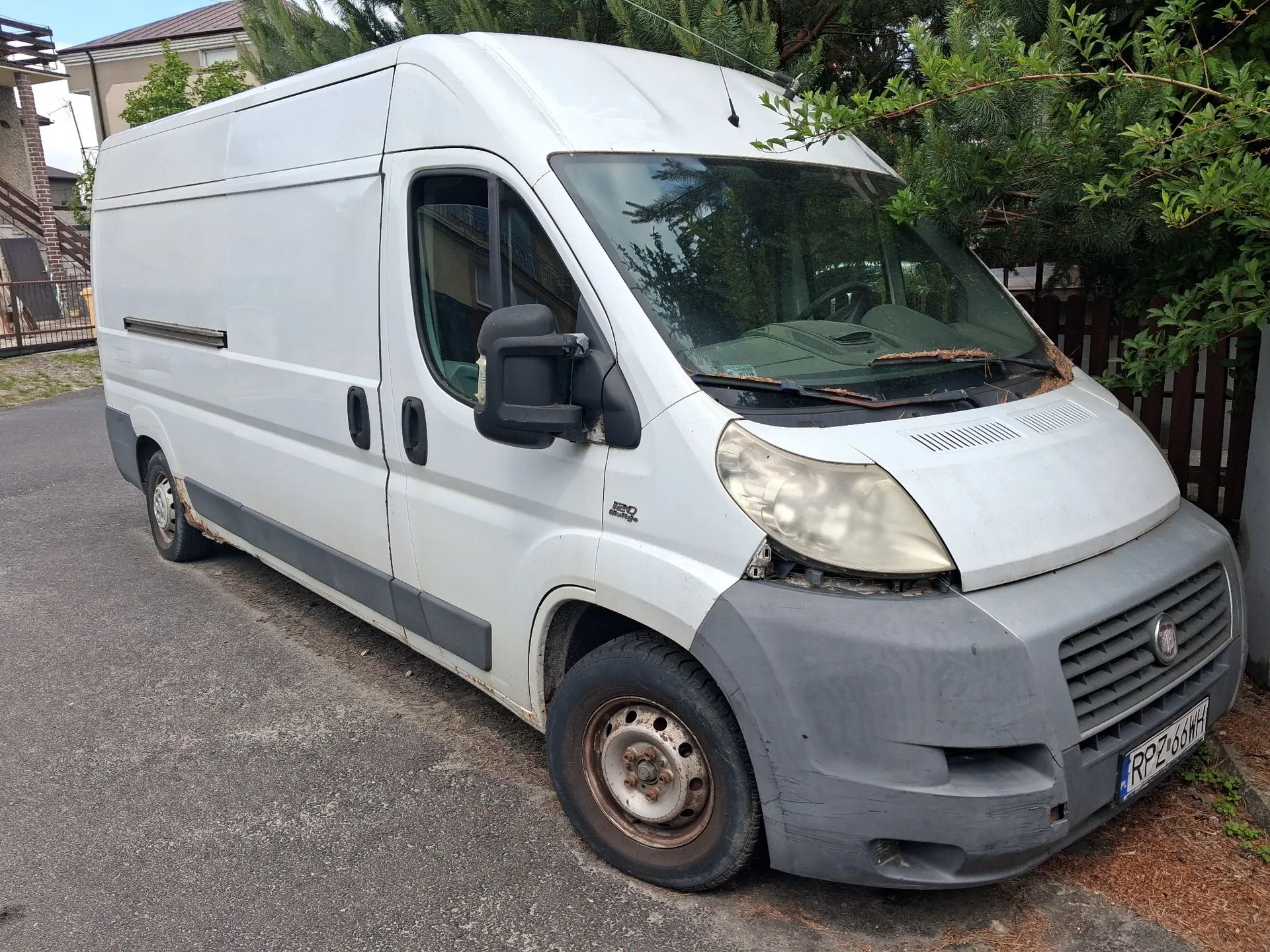 Biały furgon marki Fiat Ducato zaparkowany na asfaltowej drodze. Pojazd jest w widoku z przodu i z boku, ma szary plastikowy zderzak z przodu oraz polskie tablice rejestracyjne o numerze RPZ 66WH. W tle widoczny jest fragment budynku oraz zielone drzewa i krzewy.