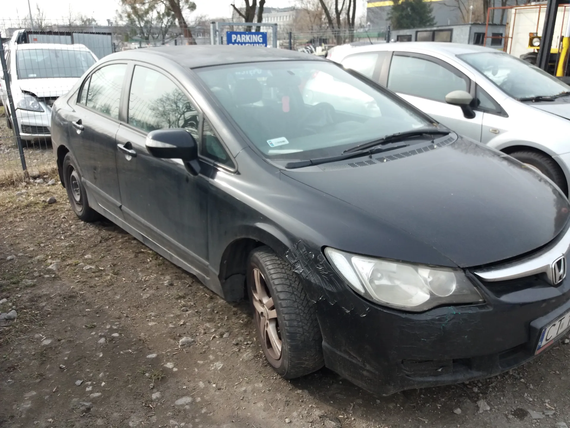 Czarny samochód osobowy marki Honda z uszkodzonym przodem, zaparkowany na nierównym terenie z ziemią i kamieniami. W tle białe samochody, w tym jeden z uszkodzonym przodem, oraz niebieski znak z białym napisem 'PARKING'. Widoczna częściowo tablica rejestracyjna z napisem 'CT'.