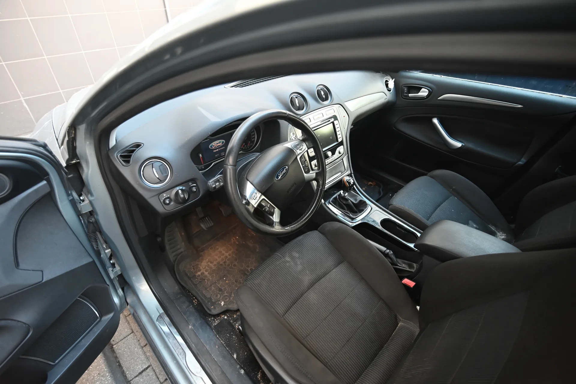 Wnętrze samochodu marki Ford z otwartą lewą przodem drzwiami. Widoczna kierownica z logo Ford, deska rozdzielcza z ekranem i przyciskami, czarne siedzenia oraz zabrudzone dywany podłogowe. W tle fragment budynku z płytkami.