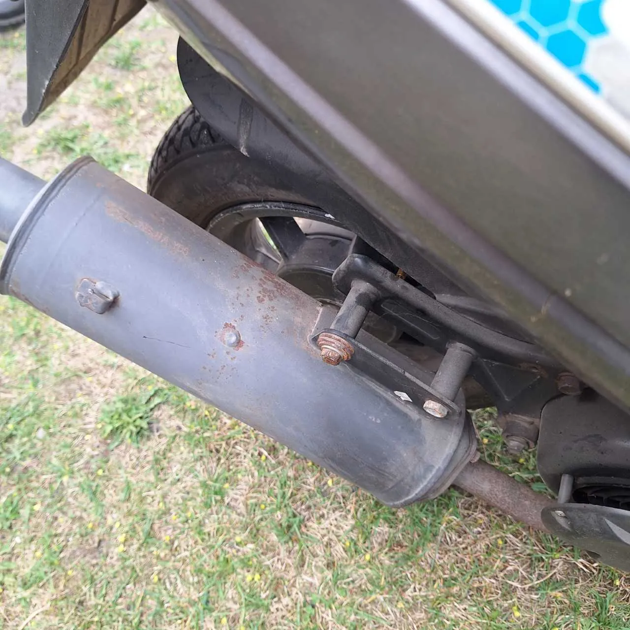 Zbliżenie na wydech motocykla z widocznymi śladami rdzy i śrubami, koło tylne oraz fragment nadwozia z niebieskim wzorem w kształcie sześciokątów. Tło stanowi trawa z żółtymi kwiatkami.