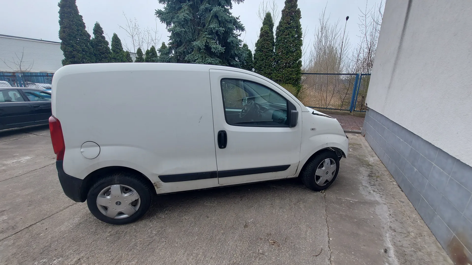 Biały furgon zaparkowany na betonowej powierzchni obok budynku z białą ścianą i szarymi płytkami na dole. W tle widoczne są drzewa iglaste, czarny samochód osobowy, błękitne ogrodzenie oraz budynek z białą elewacją. Furgon ma czarne akcenty na zderzakach i boku, srebrne felgi oraz czerwone światło tylnego. Na tle niebo jest pochmurne.