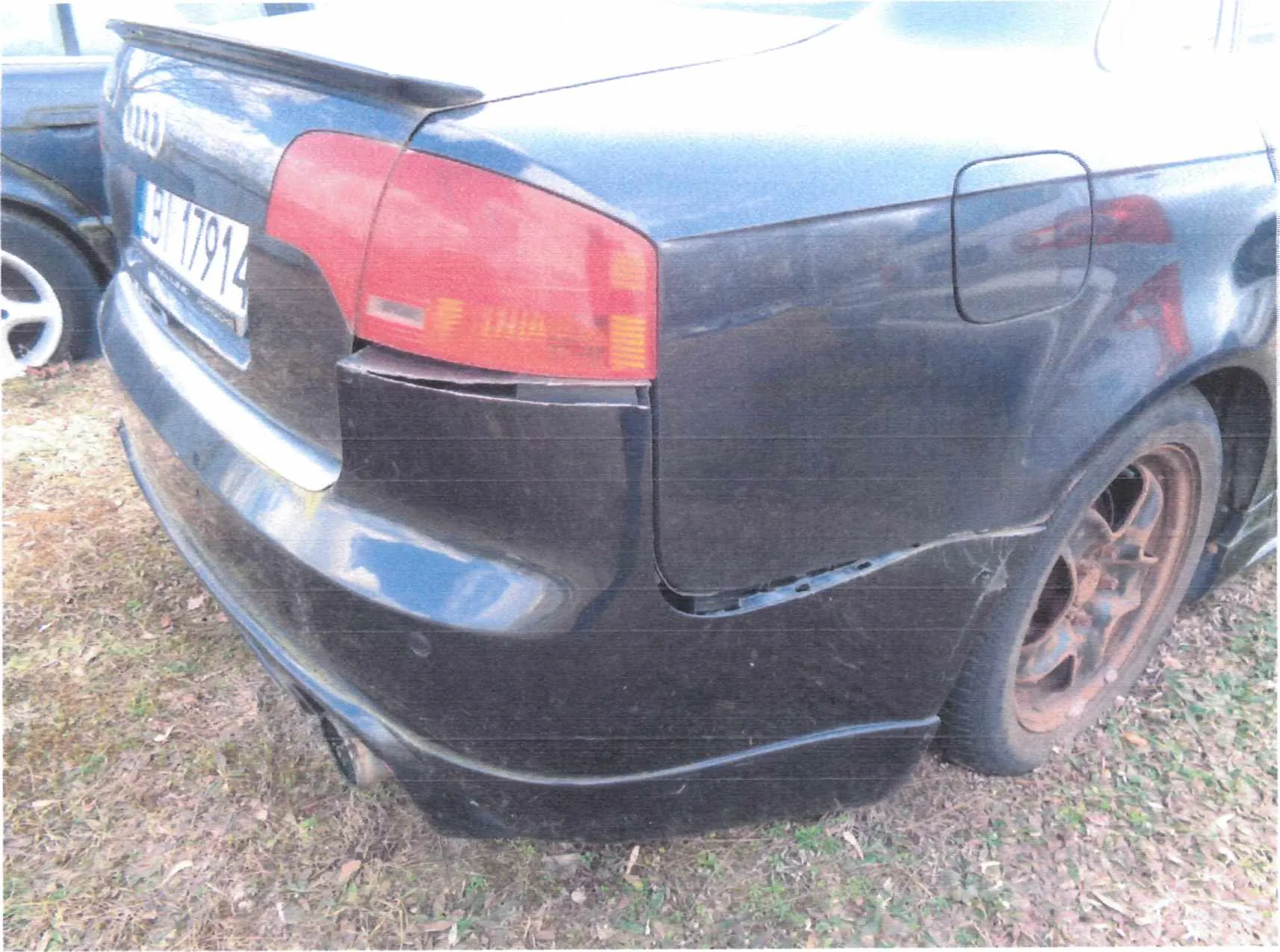 Zdjęcie przedstawia tylną część czarnego samochodu marki Audi z widocznym logo i tablicą rejestracyjną BI 17914. Samochód ma uszkodzony tylny zderzak, czerwone światło tylnego reflektora oraz zardzewiałe felgi. W tle częściowo widoczny jest inny pojazd i trawa z suchymi plamami.