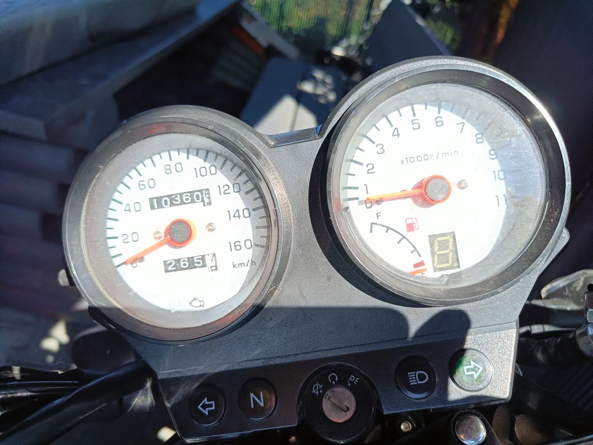 Zbliżenie na zestaw wskaźników motocyklowych. Po lewej znajduje się prędkościomierz z maksymalną wartością 160 km/h, z licznikiem przebiegu wskazującym 10360 km oraz mniejszym licznikiem 2654. Po prawej znajduje się obrotomierz z zakresem do 11 x1000 r/min oraz wskaźnikiem poziomu paliwa. Poniżej zegarów widoczny jest czarny panel z przyciskami i przełącznikami, w tym oznaczeniami 'N' (neutral) oraz ikonami świateł.