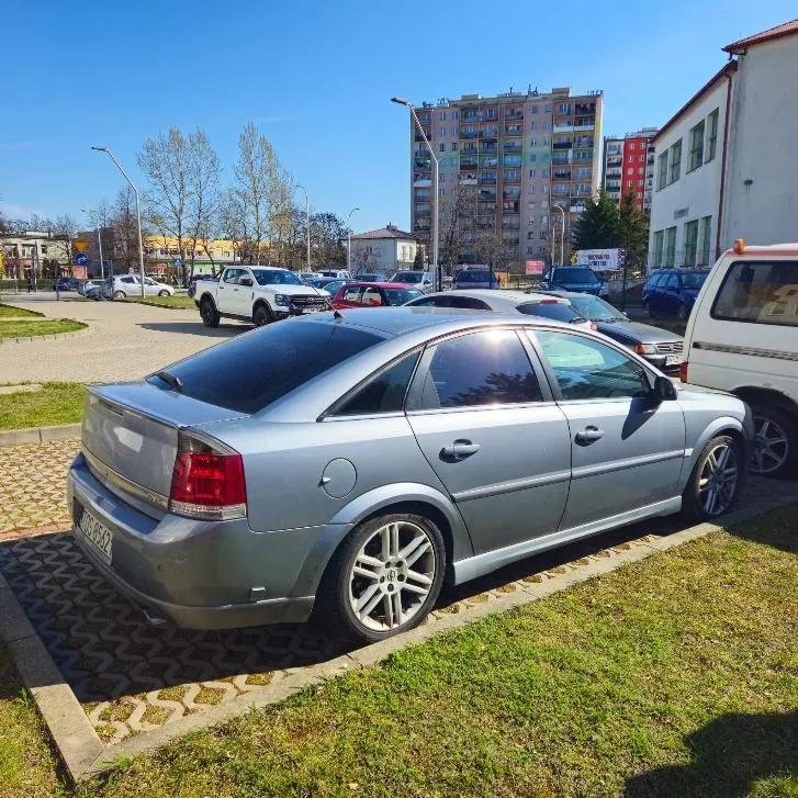Srebrny samochód osobowy typu sedan zaparkowany na trawniku obok chodnika z wzorzystą kostką. Widoczna tablica rejestracyjna HUC 8562. W tle inne pojazdy (biały pickup, czerwony samochód, biały van), wysoki blok mieszkalny, budynek z zielonymi oknami, drzewa bez liści i jasne niebo. Słoneczna pogoda, cienie wskazują na położenie słońca po prawej stronie.
